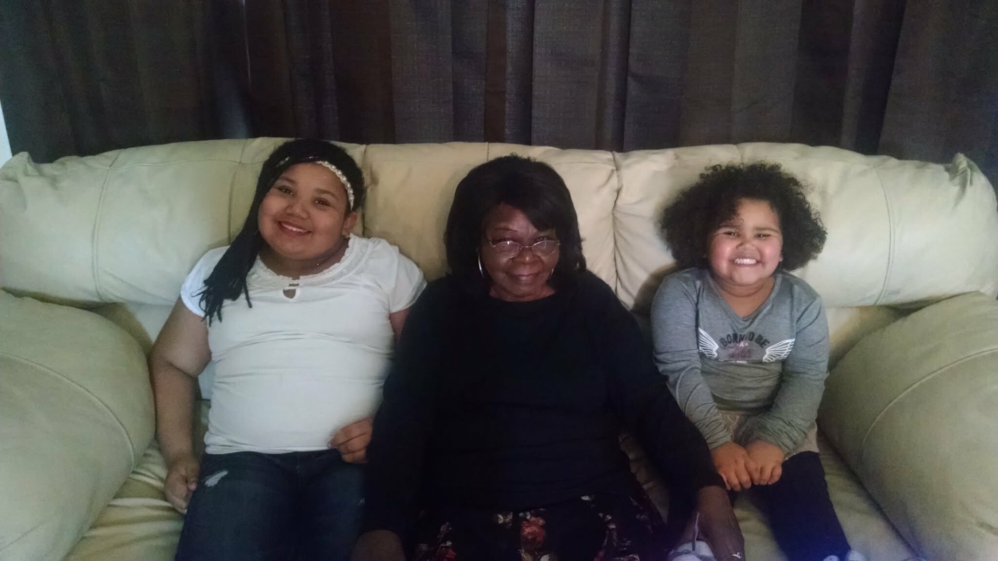 Three people sit on a beige sofa indoors: a smiling girl in white on the left, an older woman with glasses in the middle, and a smiling child with curly hair on the right.