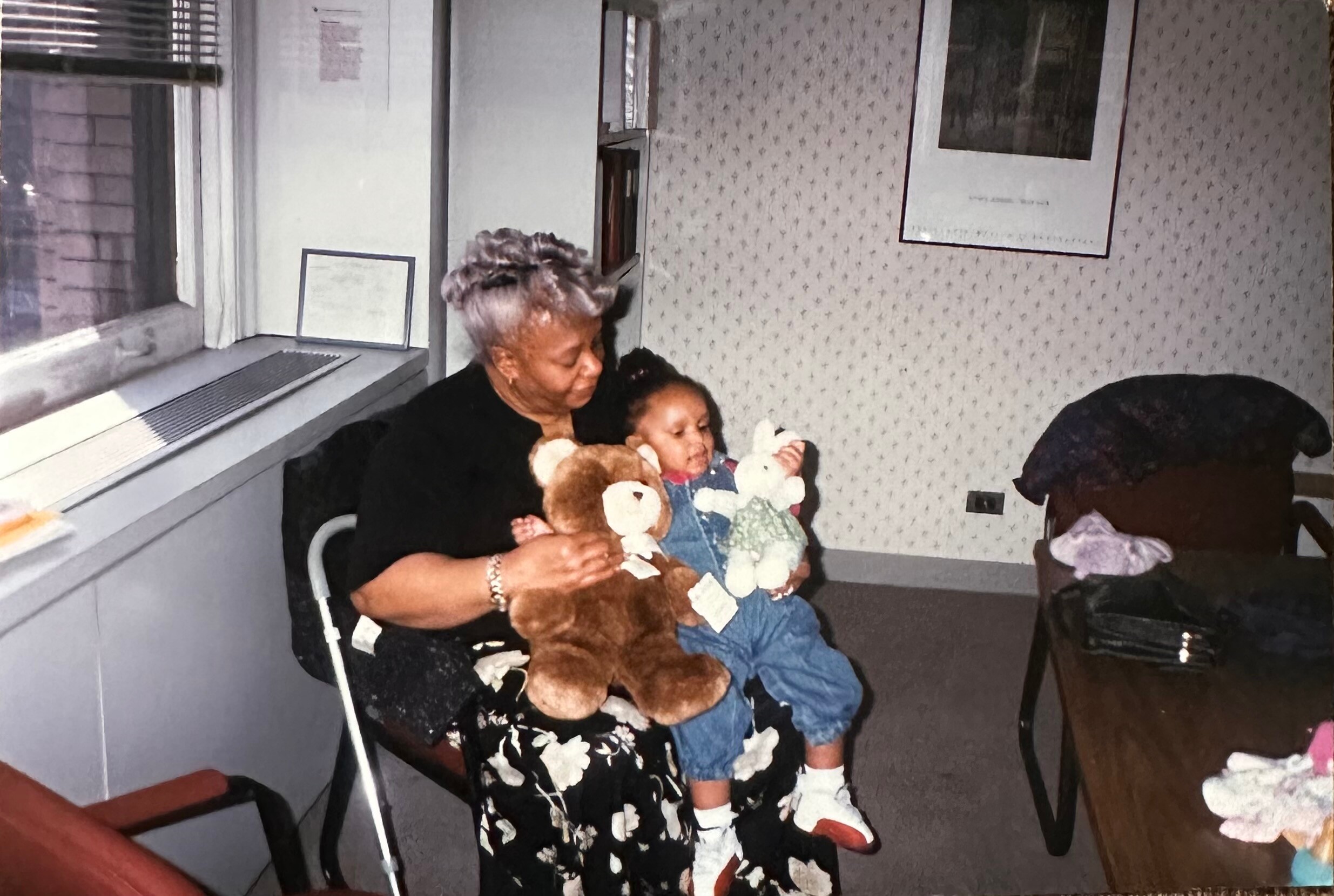 Older woman with gray hair sitting in a chair, holding a child and a stuffed bear in a cozy living room.