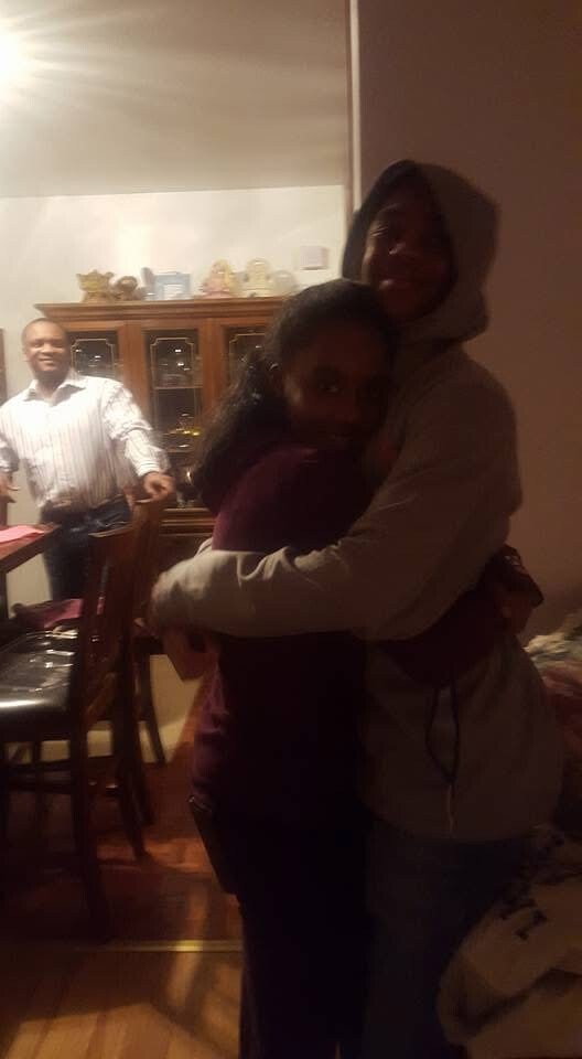 Two people share a warm hug in a dim dining room; a man stands near a table smiling in the background.