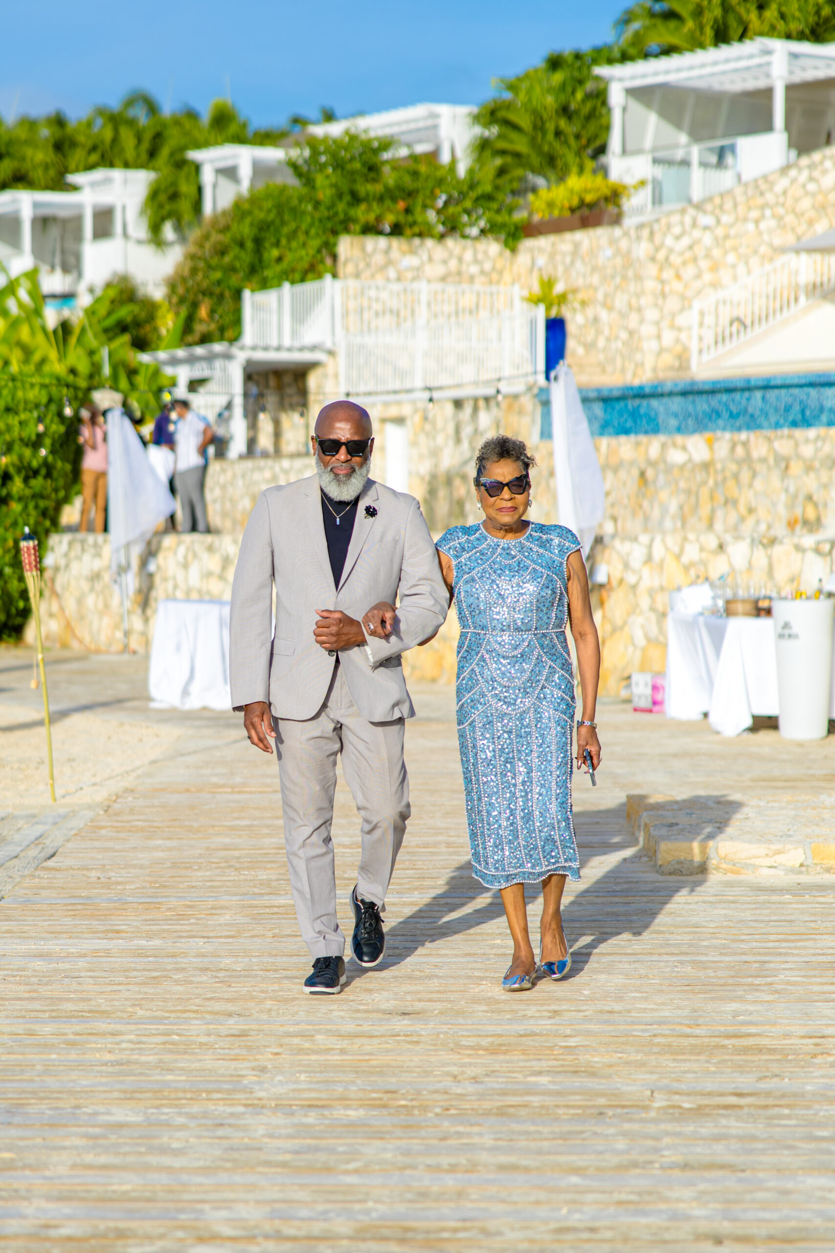 Elegant elderly couple strolling arm-in-arm along a sunny resort boardwalk, man in a light gray suit and sunglasses, woman in a blue beaded dress and sunglasses.
