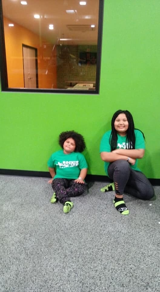 Two children sit on a gray speckled floor against a bright green wall, smiling at the camera; both wear teal shirts.