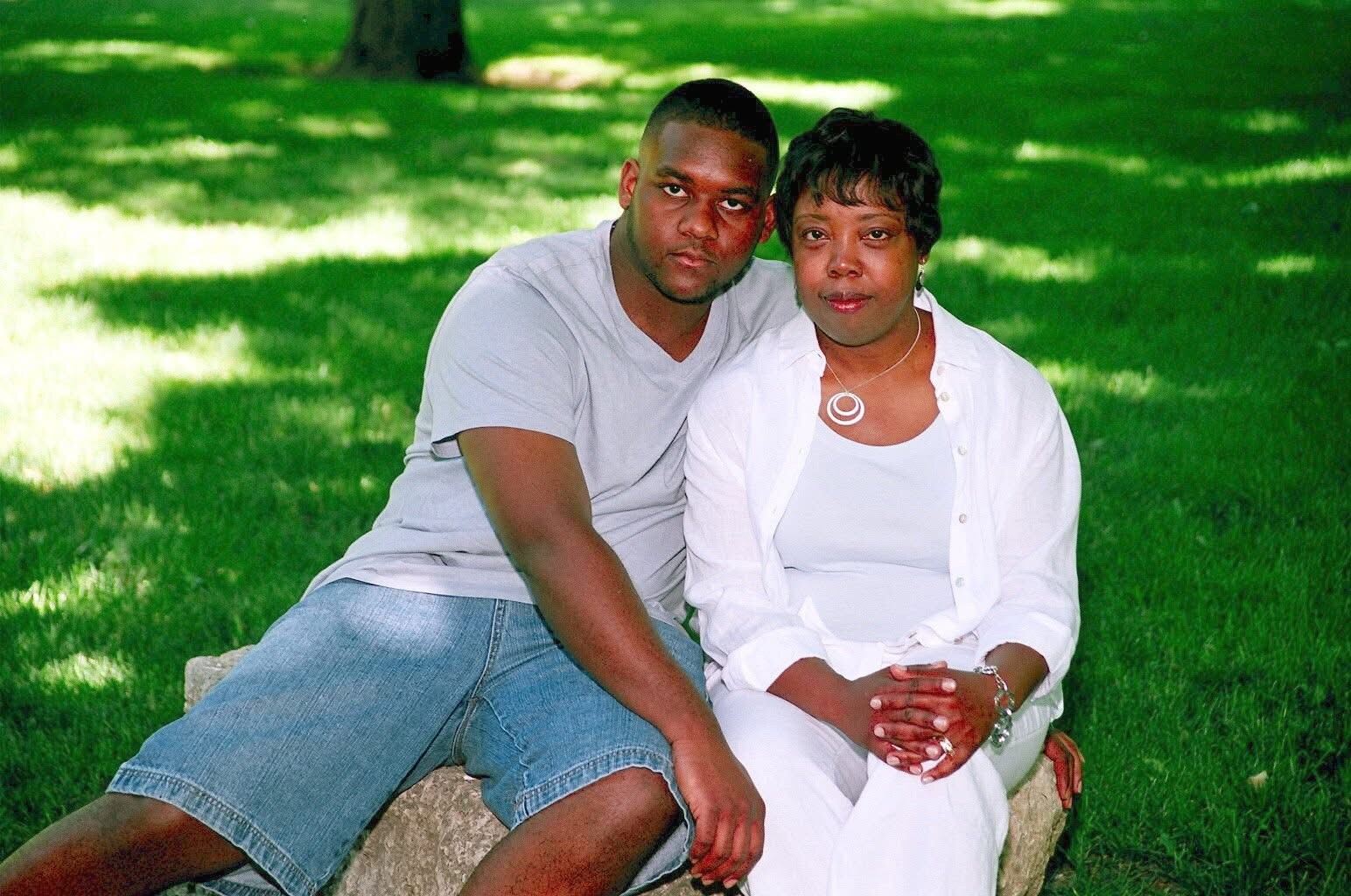 Couple sitting closely on a rock in a sunlit grassy park, looking at the camera with calm expressions. The man wears a gray T-shirt and denim shorts; the woman wears white with a white shirt layered over.