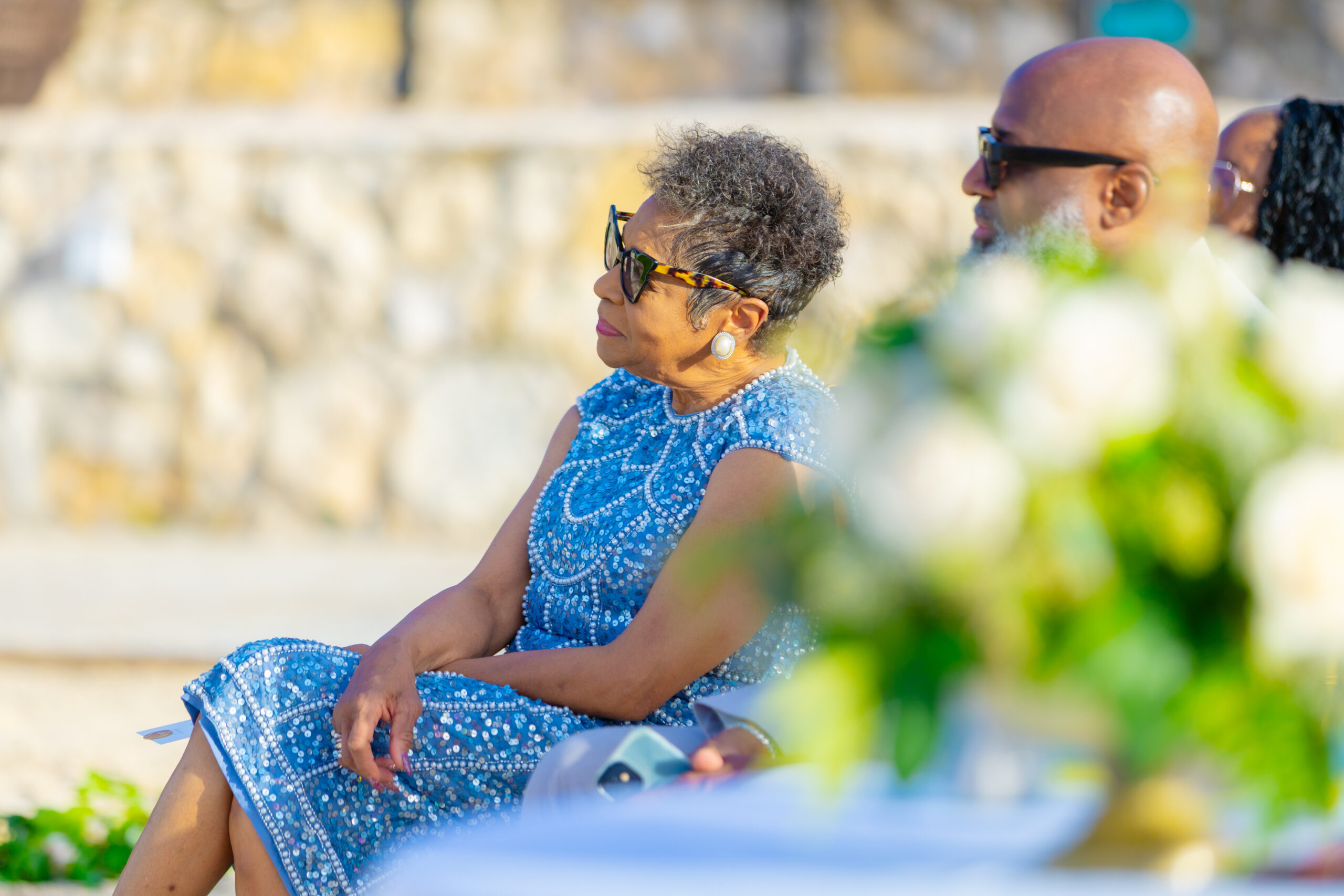 Older woman wearing a blue beaded dress and sunglasses sits outdoors, looking to the left with a calm smile.