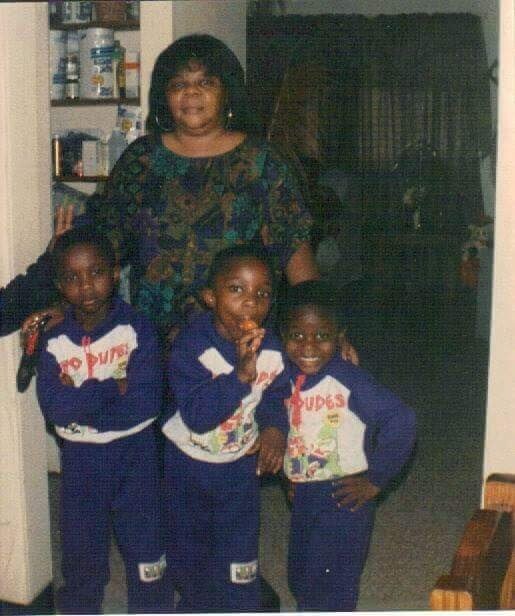 Woman stands behind three children wearing matching blue tracksuits in a home setting, shelves visible in the background.