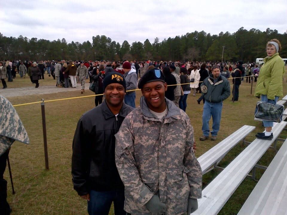 Two smiling men stand in the foreground at an outdoor event, one in camouflage and the other in a black jacket and beret, with a large crowd and bleachers behind them.