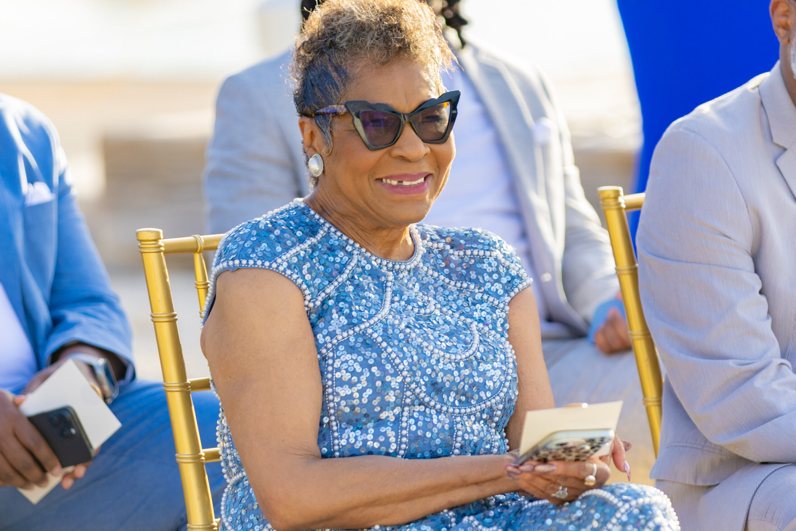 Smiling older woman in a blue sequined dress and cat-eye sunglasses, sitting outdoors and holding a smartphone.