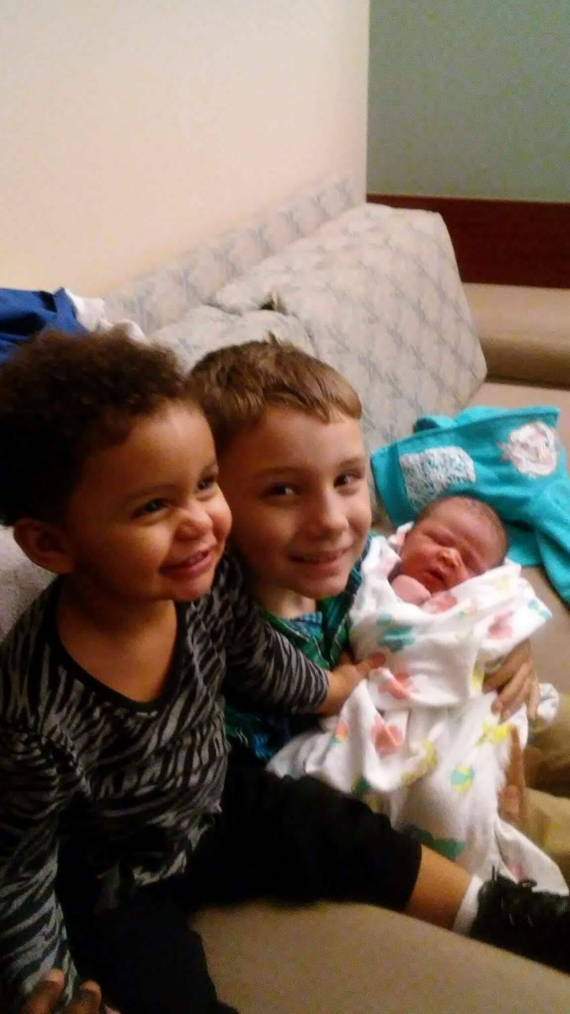 Two smiling children cradle a newborn baby wrapped in a pastel blanket on a couch indoors.