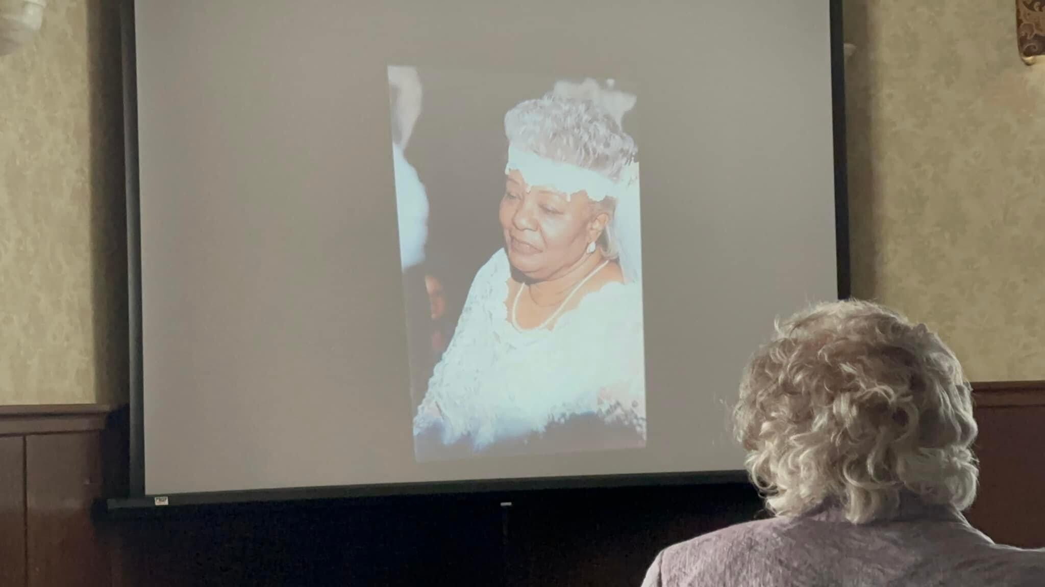 Projection screen shows a portrait of an elderly woman in a white dress and hat; audience member’s head visible in the foreground.