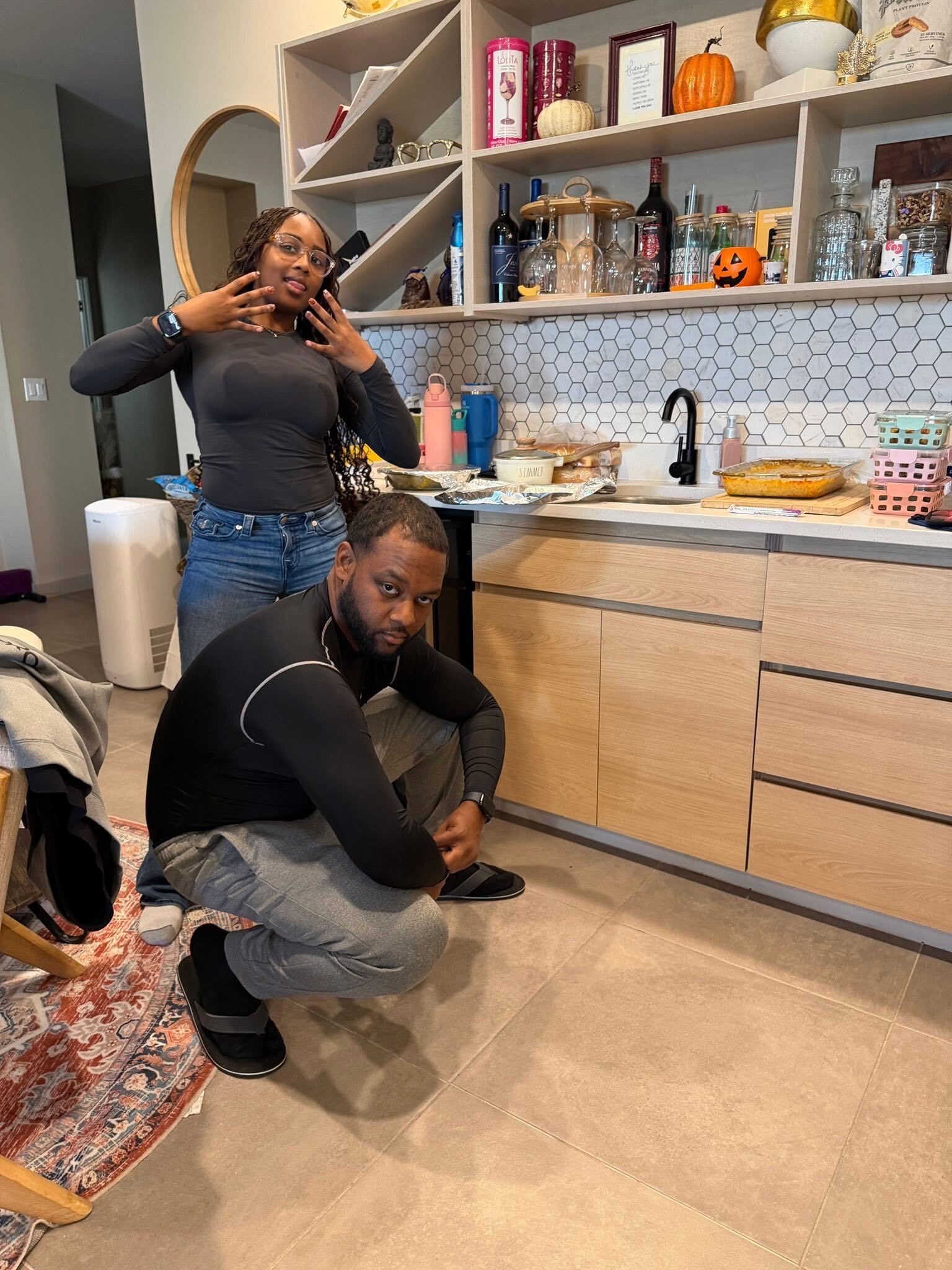 Woman stands behind a kneeling man in a modern kitchen, hands raised near her face as they pose for the photo.