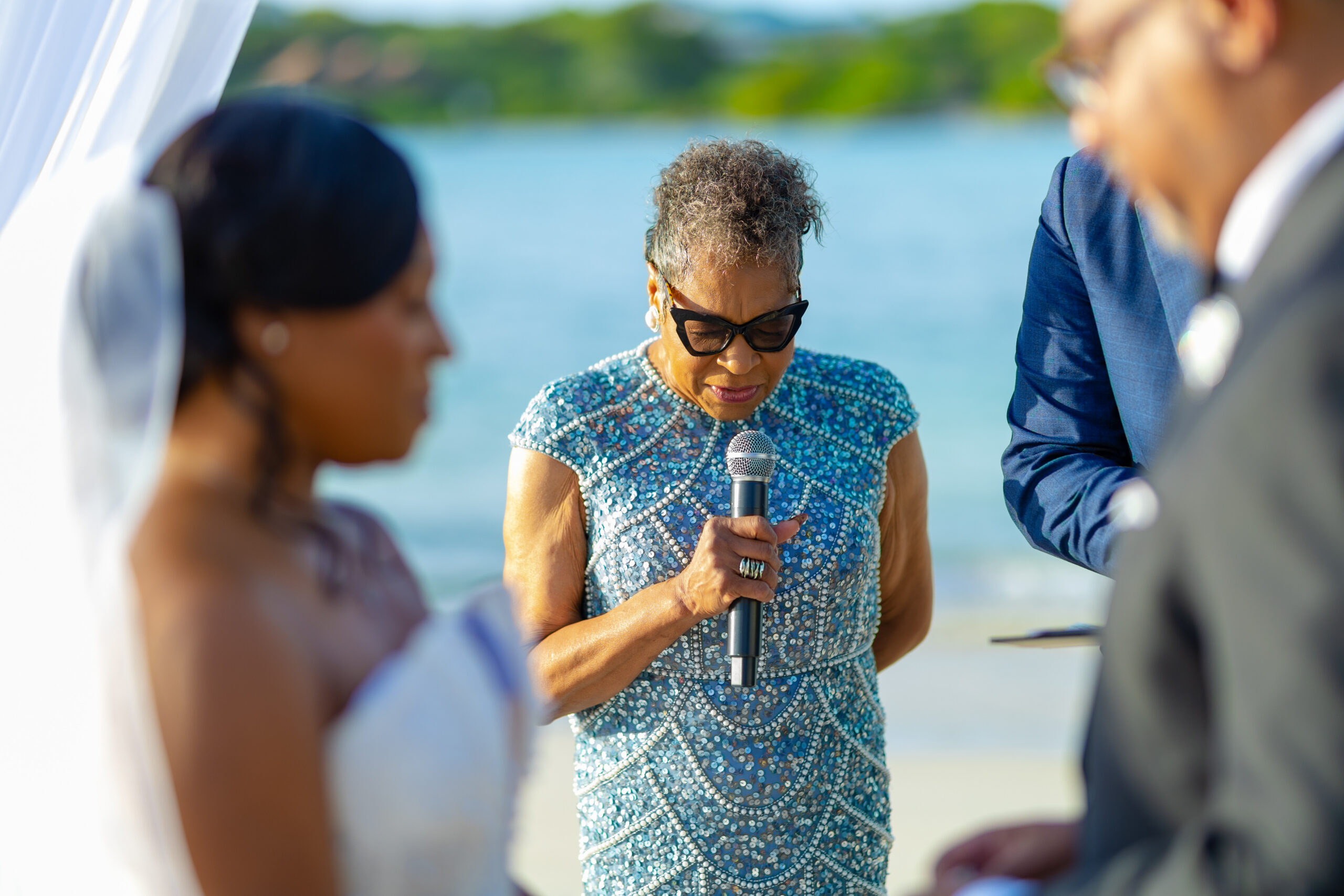 Older woman in a shimmering blue dress holding a microphone, speaking at a beach wedding as guests listen by a sunny shoreline