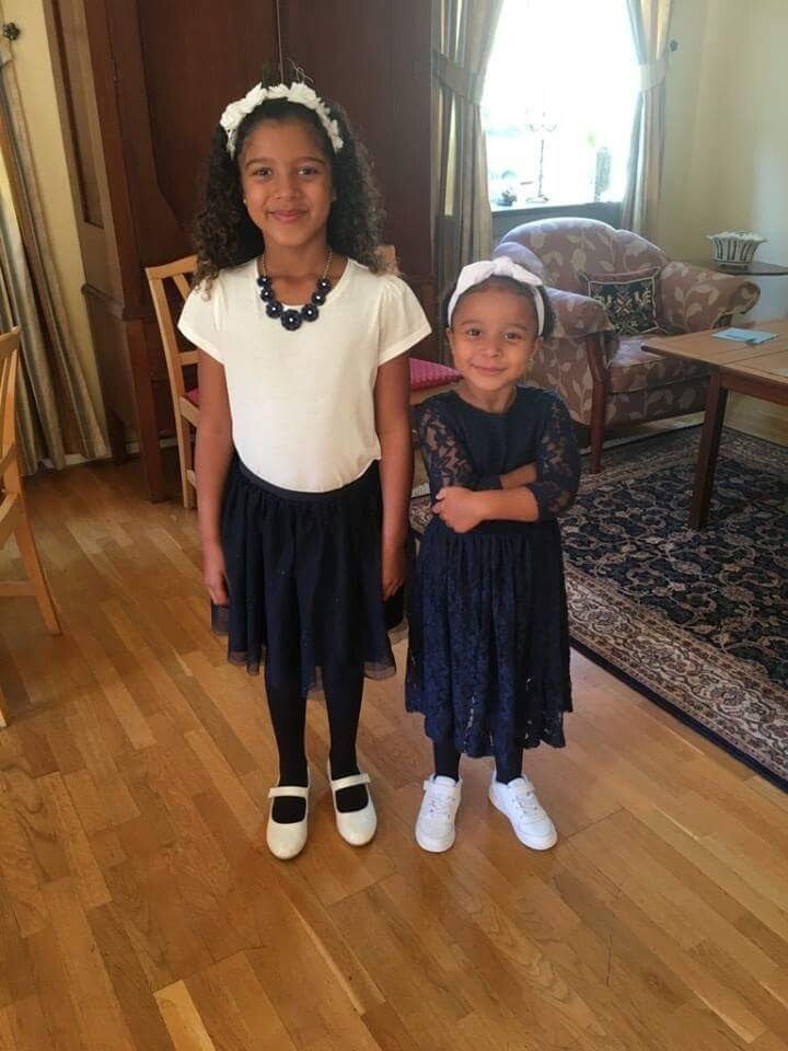 Two girls stand in a living room smiling at the camera: the older girl on the left wears a white top, navy skirt, white shoes, and a dark necklace; the younger girl on the right wears a navy lace dress with a white headband and white sneakers.
