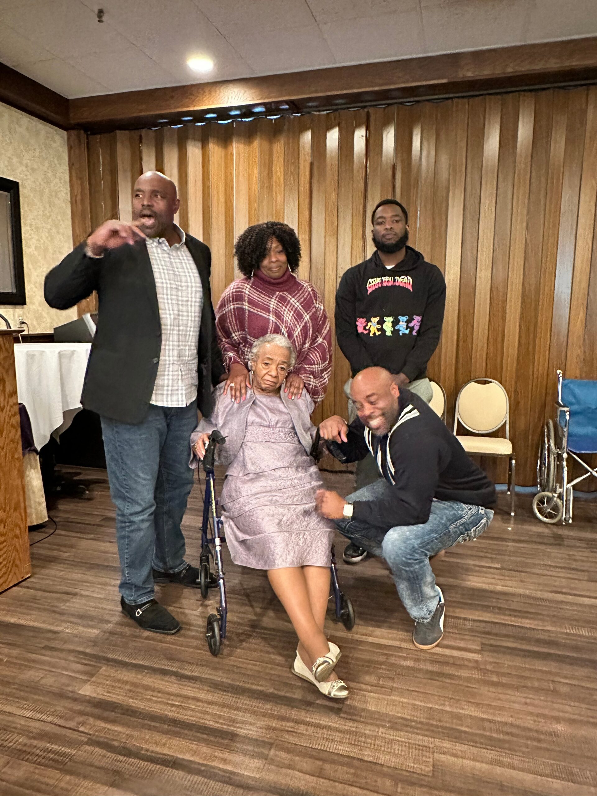 Elderly woman in a wheelchair surrounded by four adults indoors, posing for a group photo; wood-paneled wall in background on stage-like setting with chairs nearby.