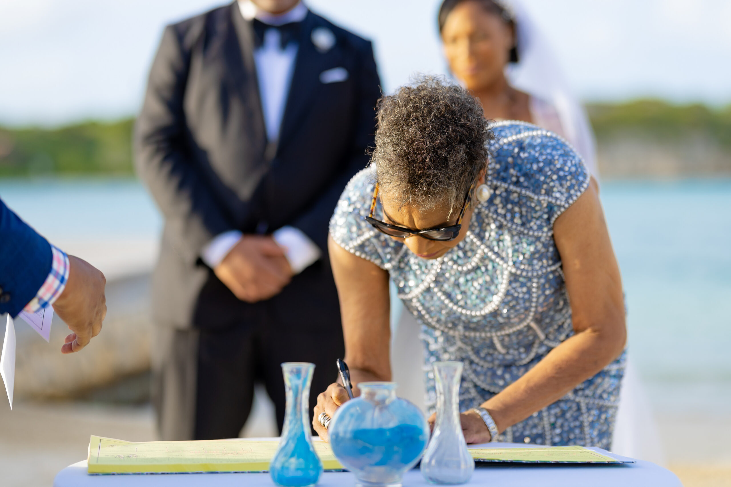 Outdoor wedding scene: a woman in a beaded dress signs a document at a table with blue glass vases in the foreground; a couple stands blurred in the background.