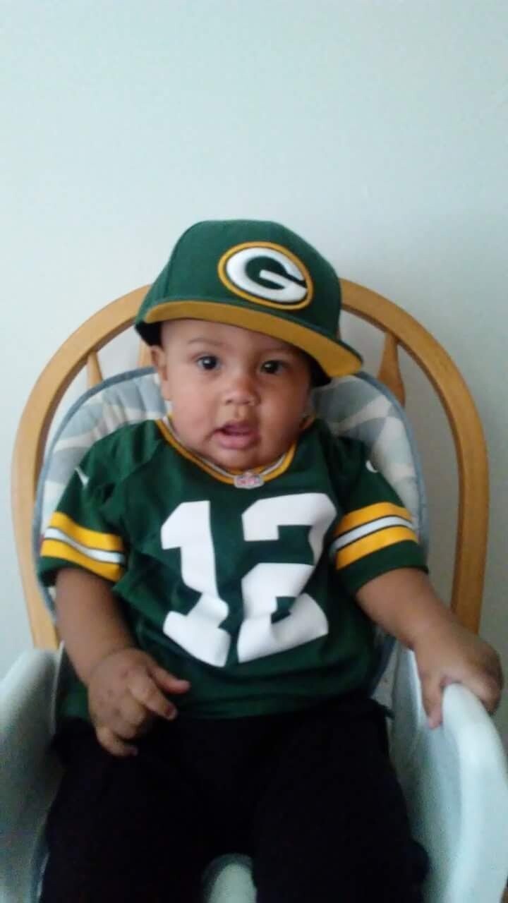 Baby in a Packers jersey (number 12) and green cap, seated in a wooden high chair indoors, looking at the camera.
