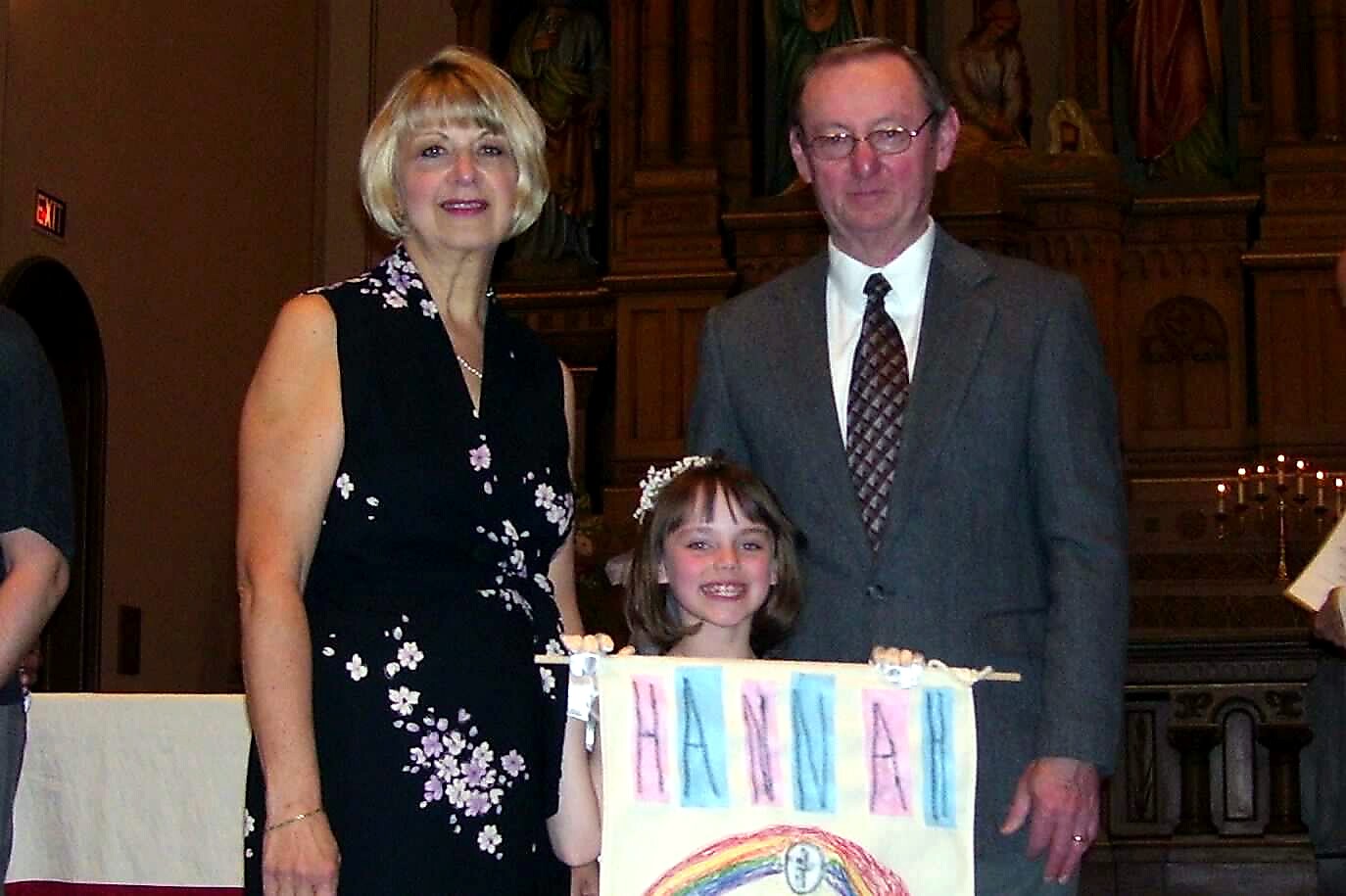Young girl with a flower headband stands beside a woman in a black floral dress and a man in a gray suit at a church, holding a rainbow banner that reads HANNAH.