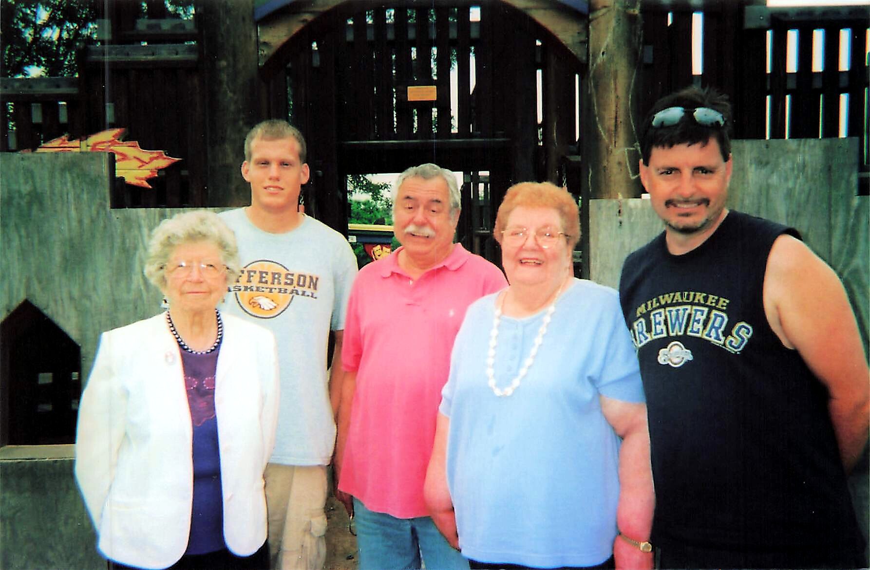 Five people posing for a photo outdoors in front of a wooden fence: an elderly woman in a white blazer, a young man in a gray T‑shirt, a man in a pink polo, an elderly woman in a light blue top, and a man in a Milwaukee Brewers sleeveless shirt with sunglasses on his head.