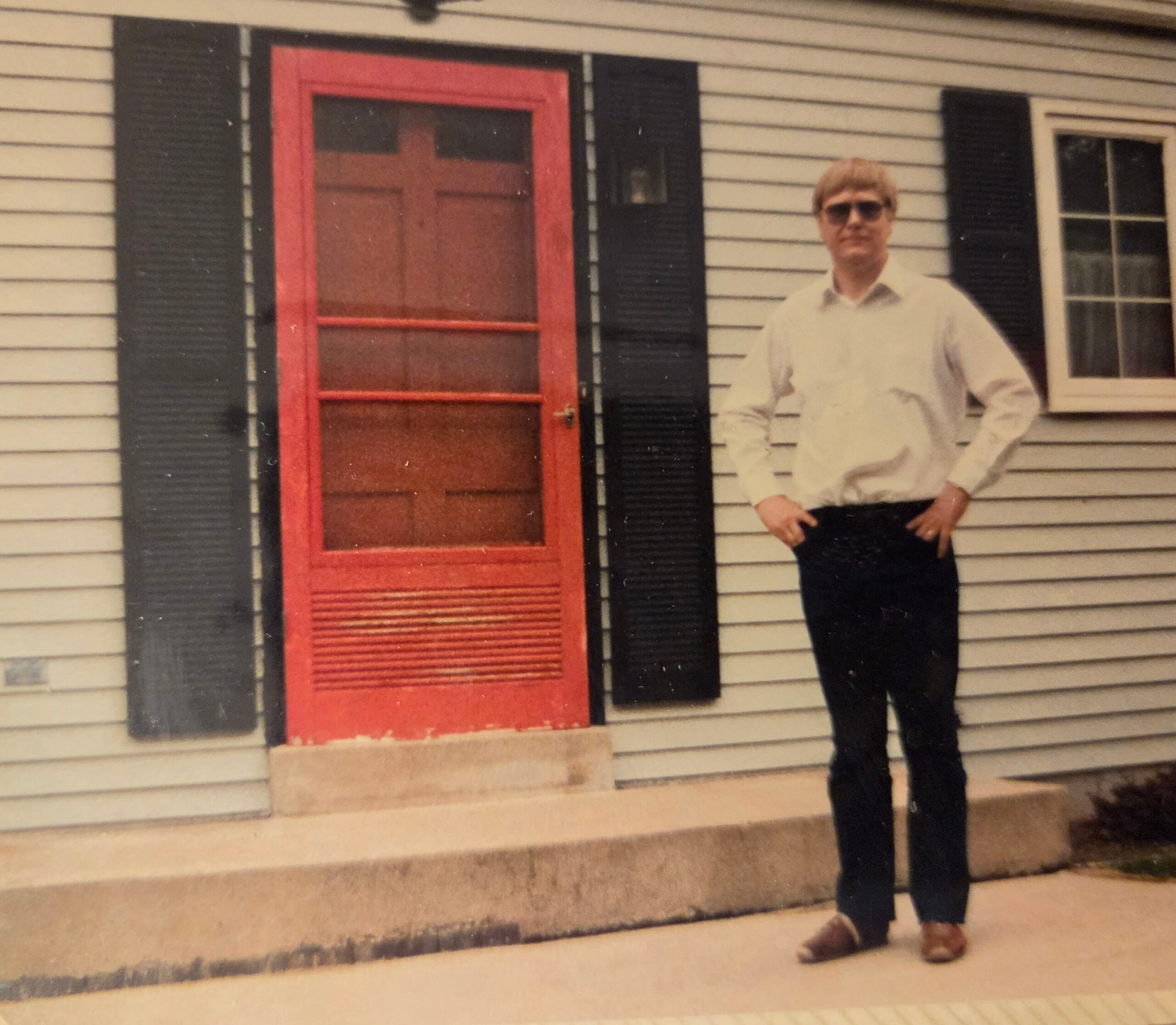 Man in a light shirt and sunglasses standing on the front steps beside a red door of a white-siding house with black shutters.