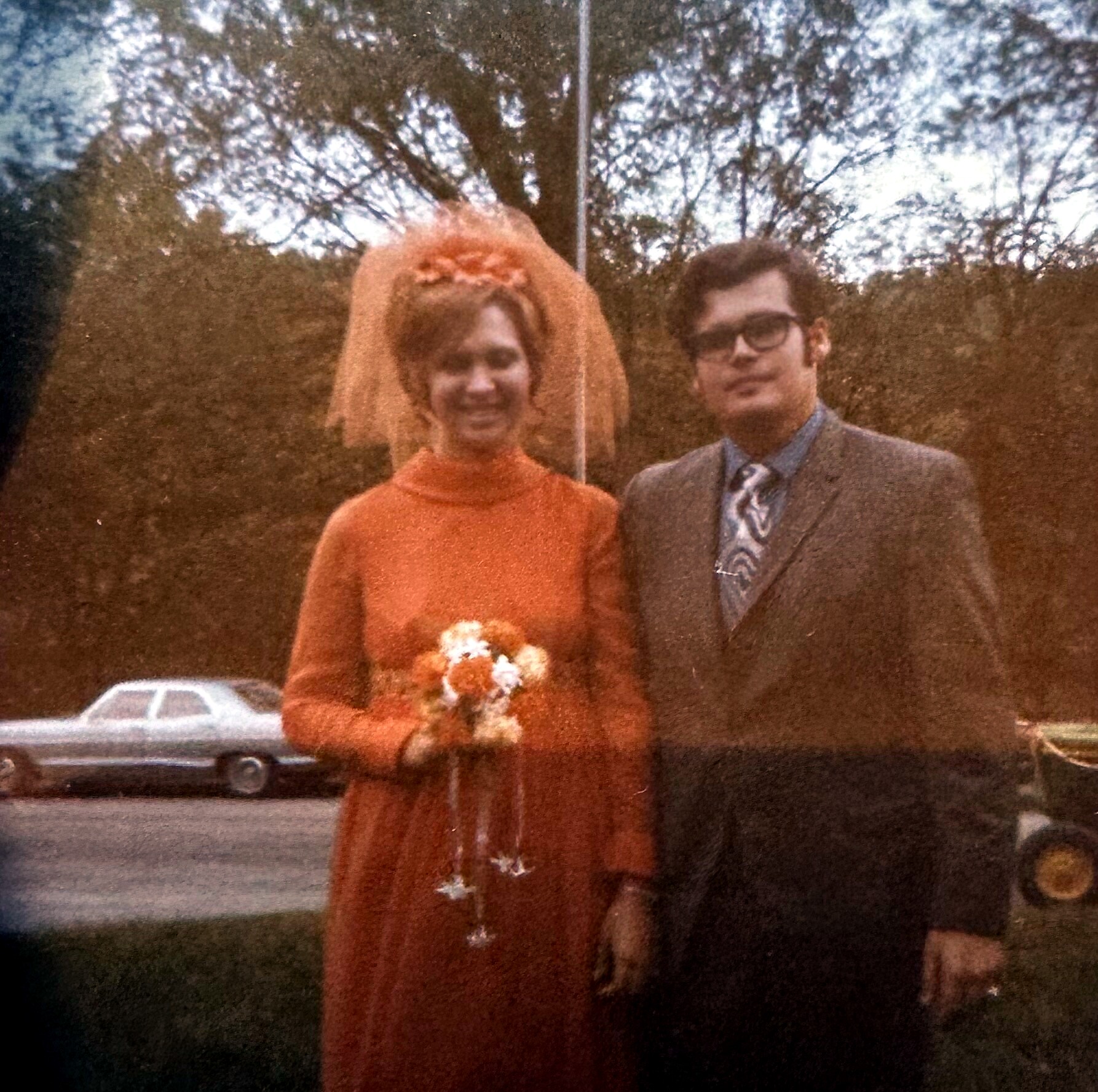 Bride in an orange dress with a veil and bouquet stands next to a groom in a suit outdoors, smiling.
