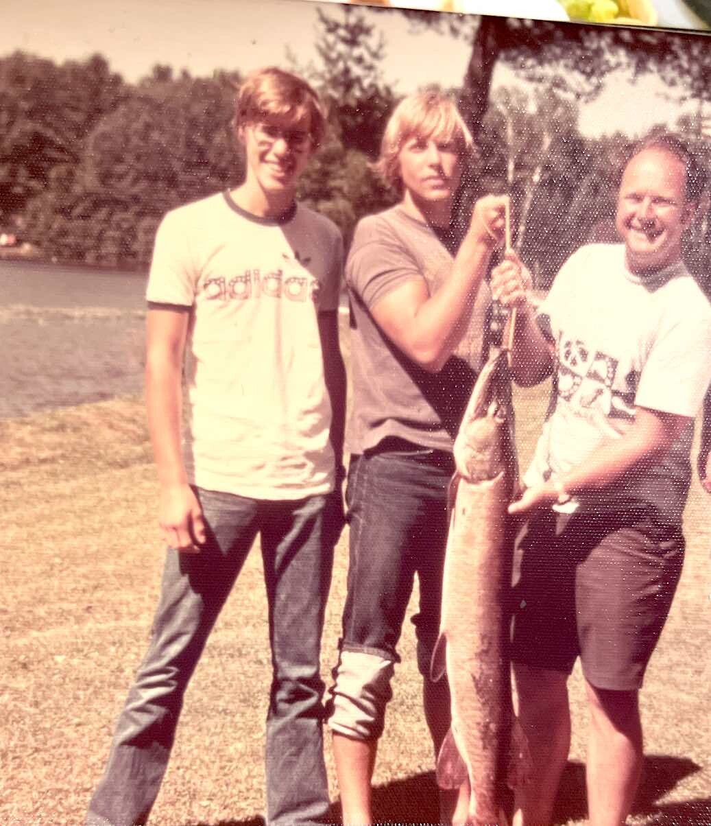 Three friends posing outdoors with a very large fish they just caught at a park/lake.