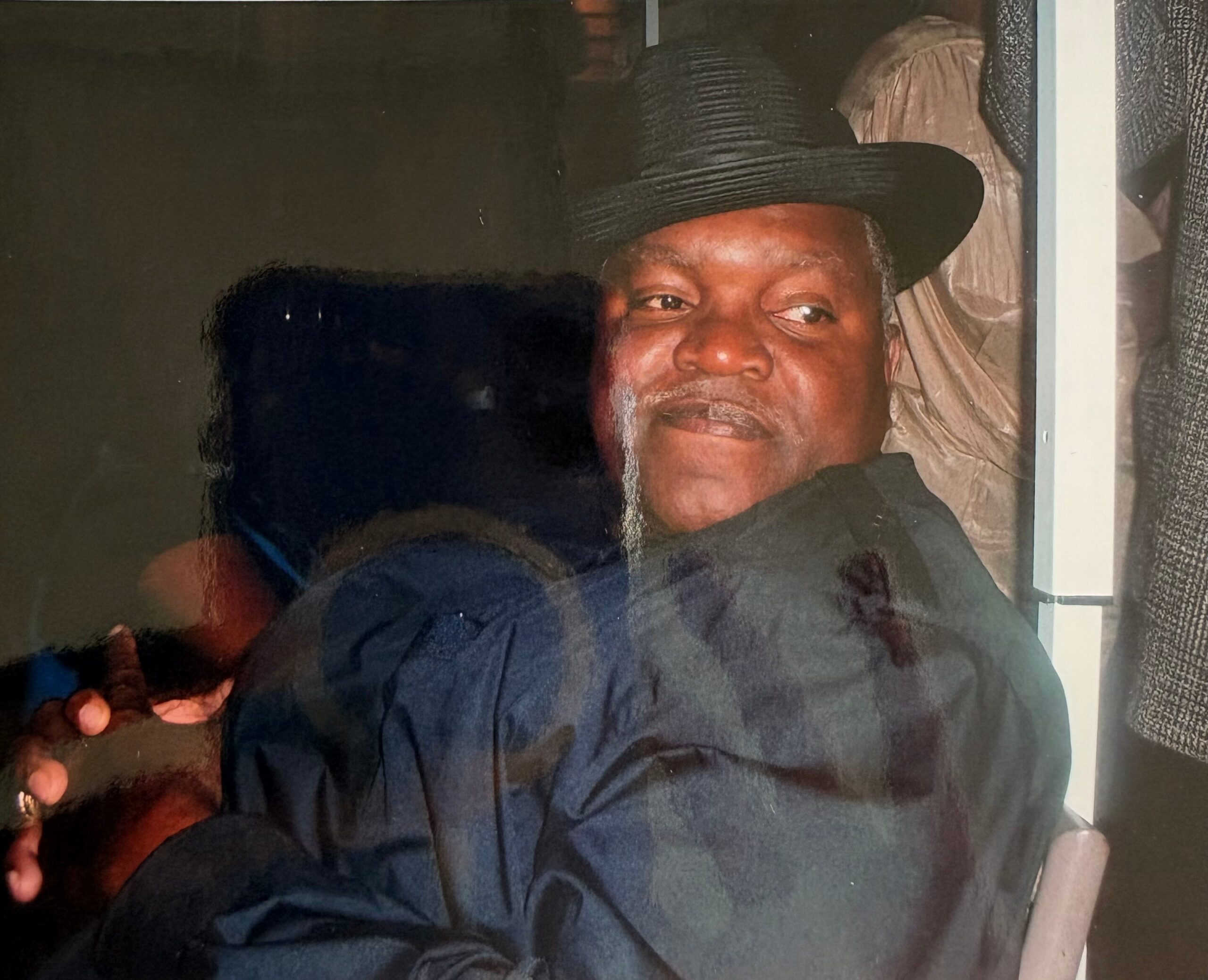 Man wearing a wide-brim black hat and dark clothing, smiling softly, in a dim indoor setting with a window behind him.