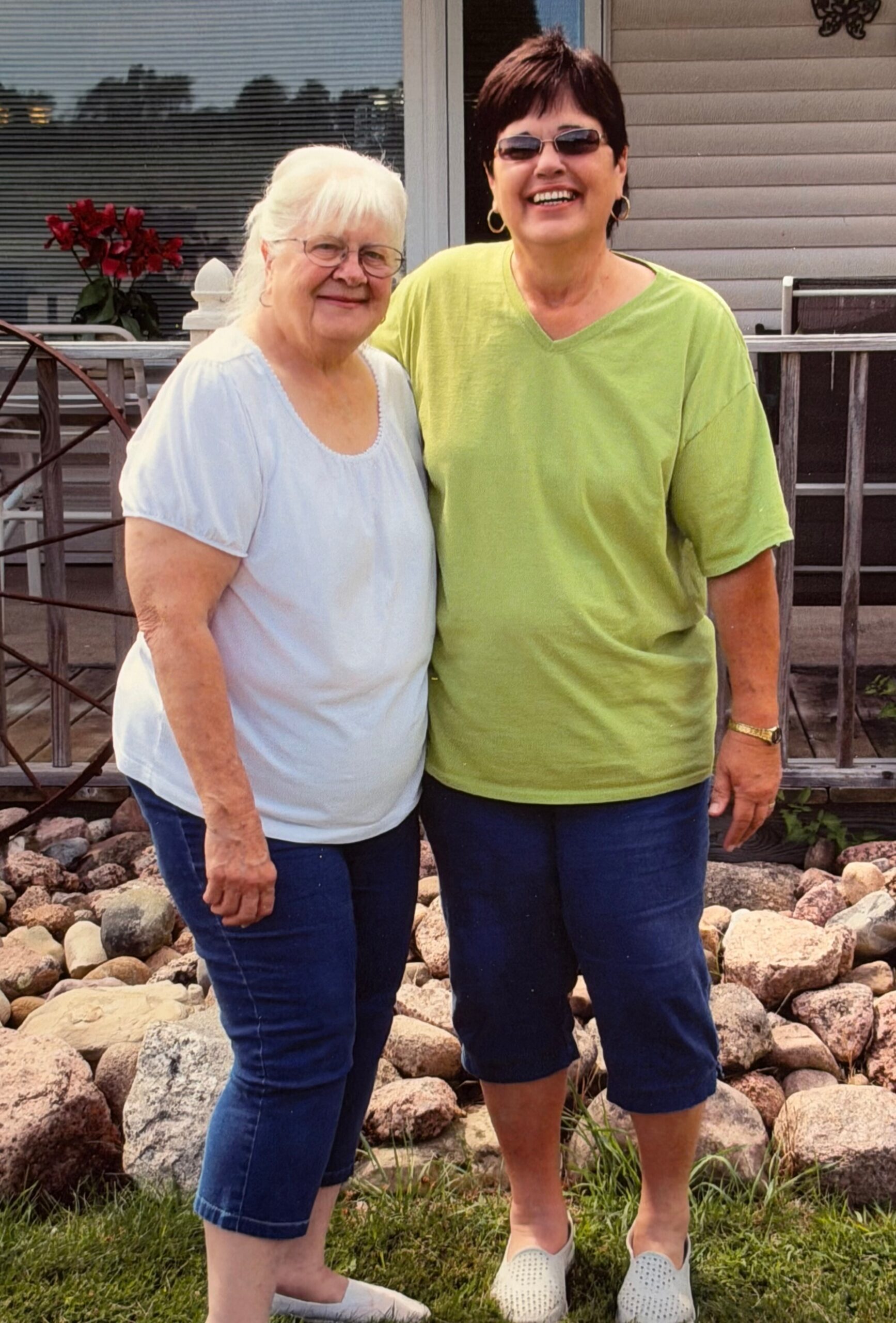 Two women stand together on a rocky lawn, smiling at the camera; one in a light blue blouse, the other in a lime green shirt and sunglasses.