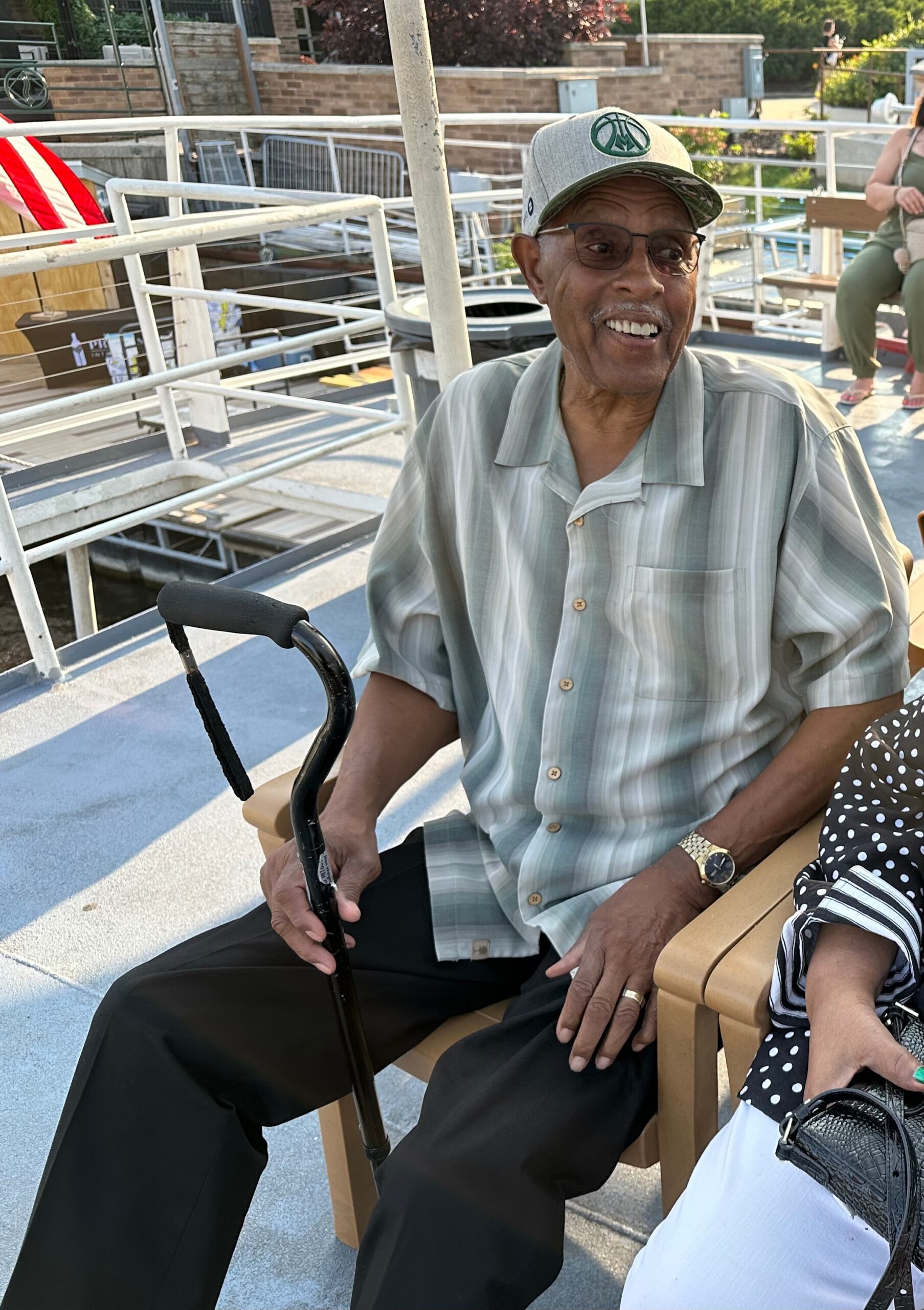 Smiling elderly man wearing a light striped shirt and cap, sitting on a wooden bench with a walking cane in his hand on an outdoor deck.