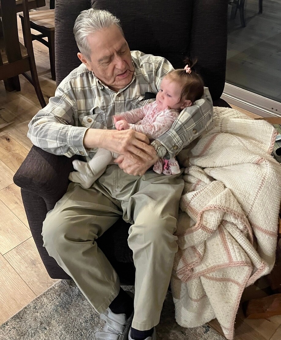 Grandfather in a plaid shirt sits in a dark armchair, cradling a baby wrapped in a blanket on his lap in a cozy living room.