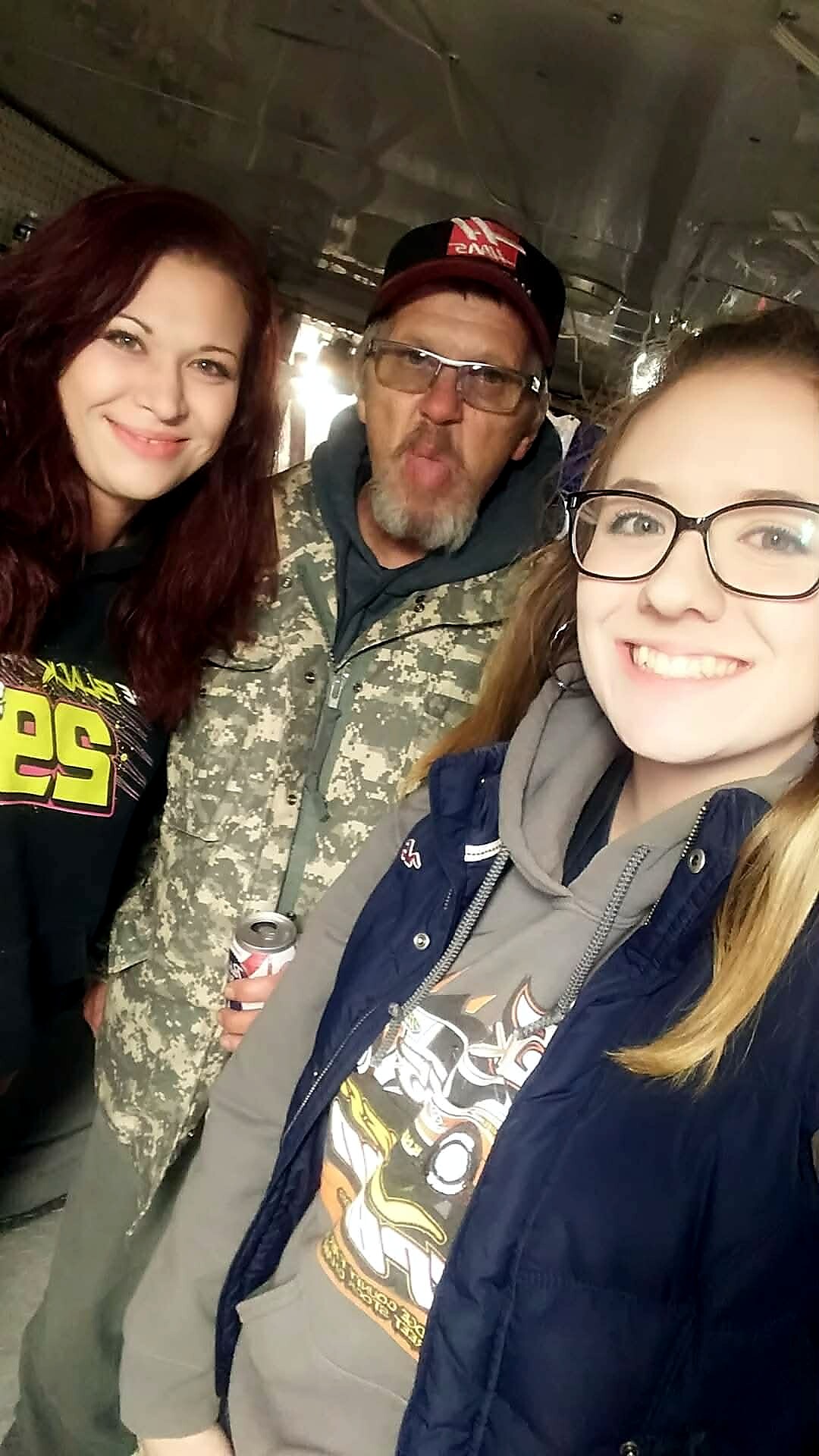 Three friends taking a casual selfie indoors: man in camo jacket center making a puckered face, woman with red hair left, woman with glasses right holding a drink can nearby.