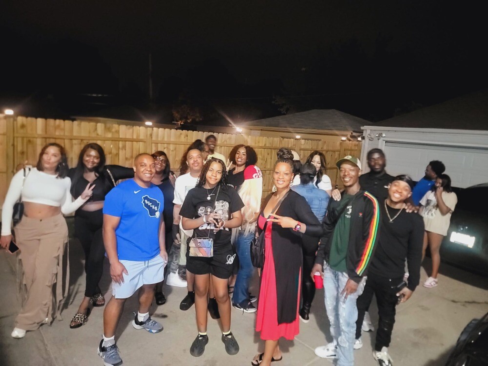 Group of friends posing together in a driveway at night, smiling and making peace signs. A wooden fence and string lights are in the background.