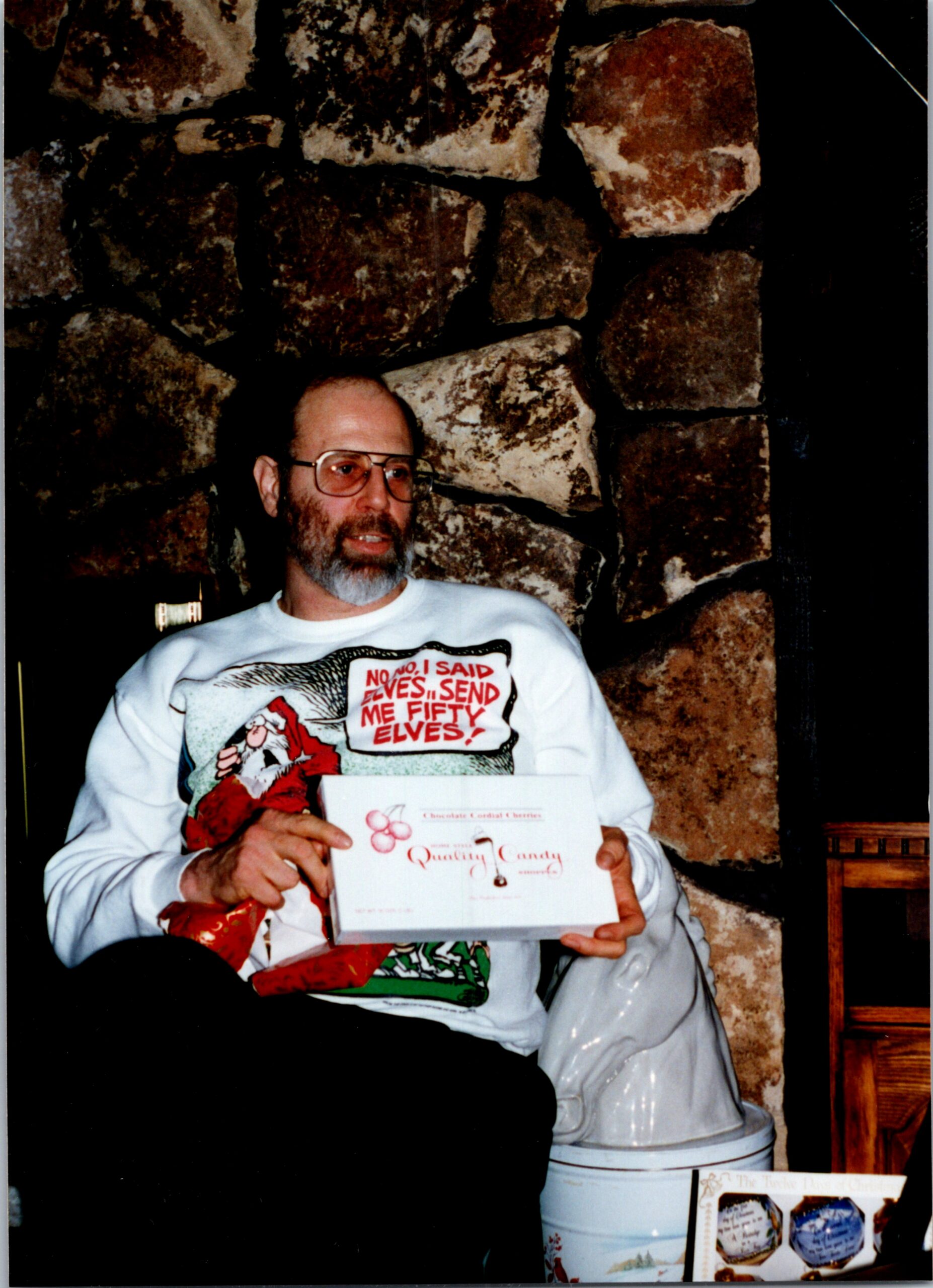 Bearded man in a white Santa-themed shirt holds a gift box while seated against a large stone wall backdrop.”,