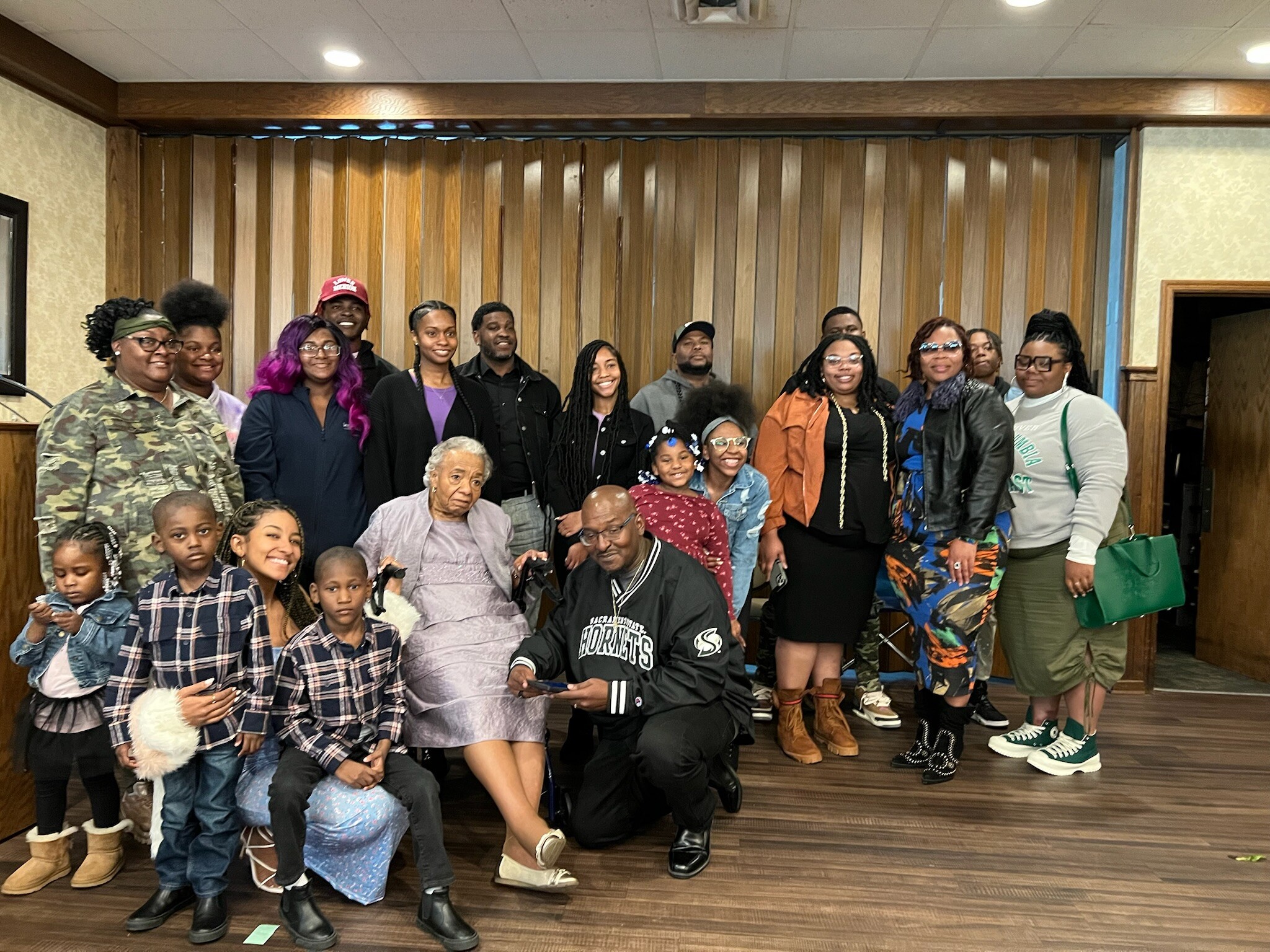 Group of diverse adults and children in colorful costumes posing for a photo indoors in a room with wood paneling.