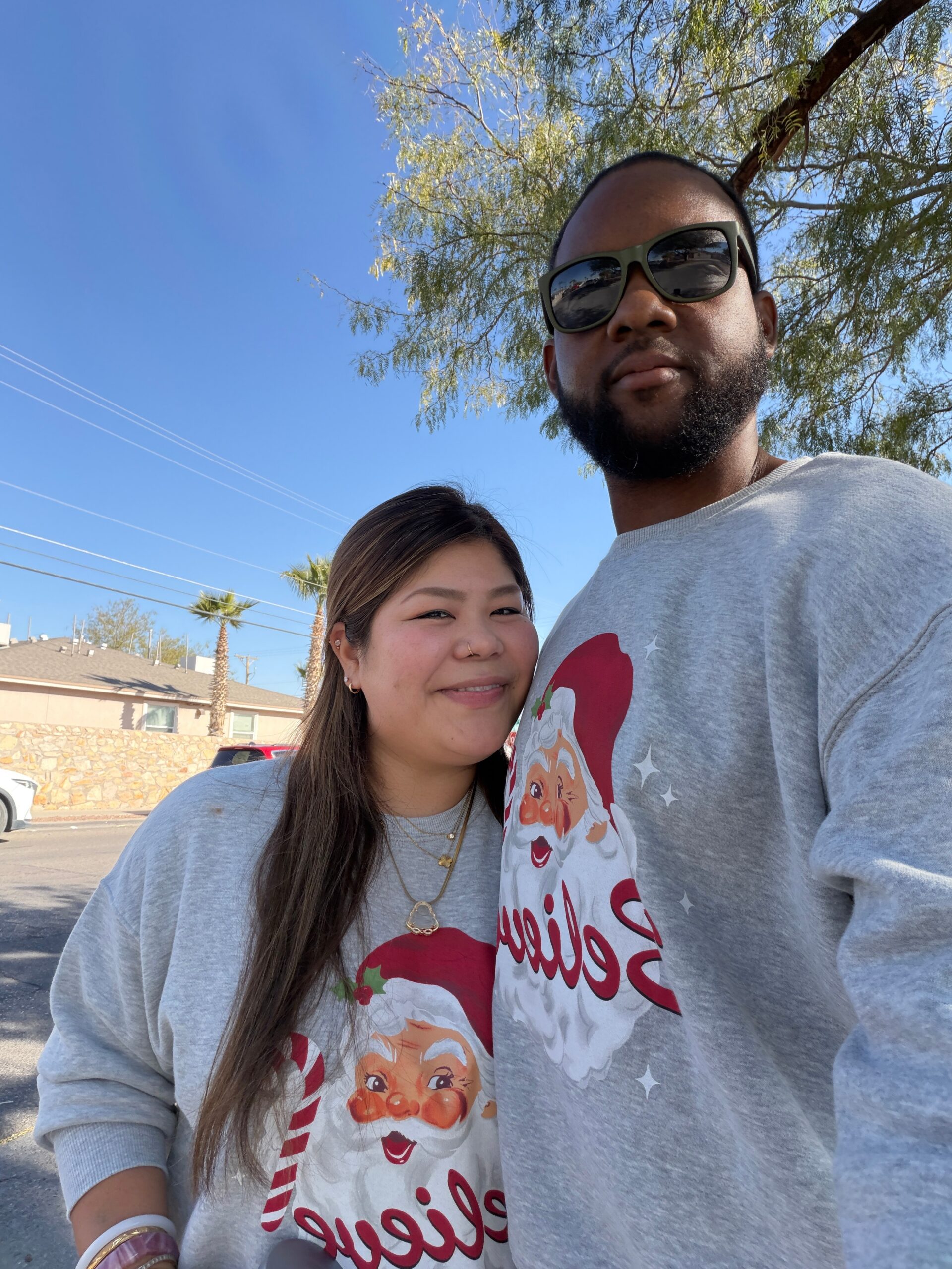 Man and woman posing for a selfie outdoors on a sunny day, both wearing gray Christmas sweaters with Santa Claus graphics.