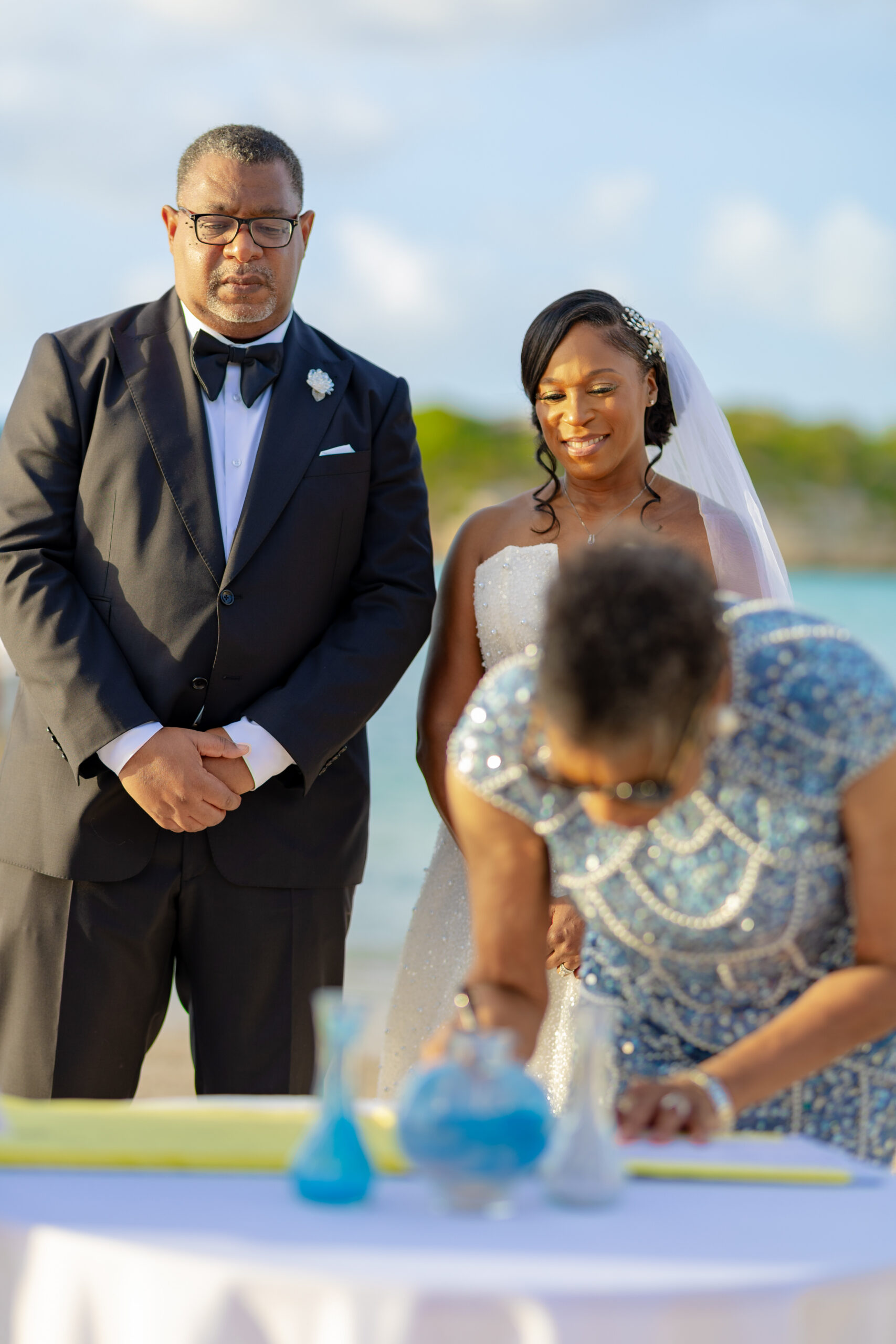 Bride in a white gown and groom in a black tuxedo stand outdoors, smiling as they attend a table with blue decorations during a wedding.
