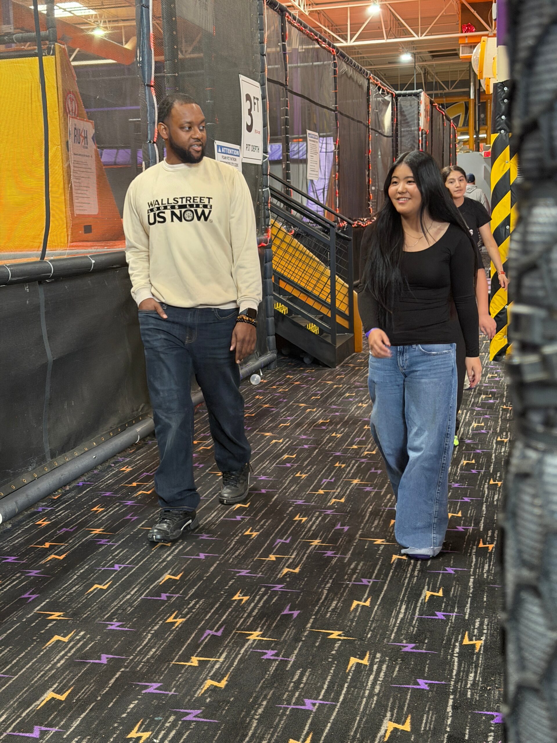 Man in beige sweatshirt and woman in black top walk through an indoor trampoline park corridor with safety nets and padded walls.