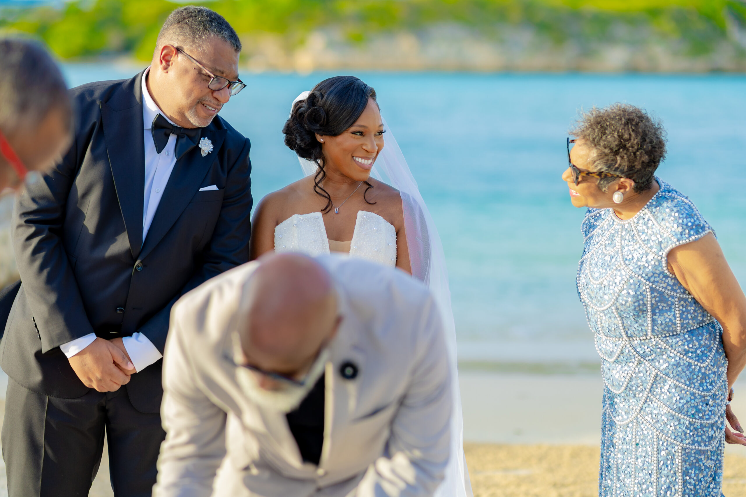 Bride in a white strapless dress and veil smiles beside a man in a black tux on a beach by the water; others nearby in formal attire admire the moment.
