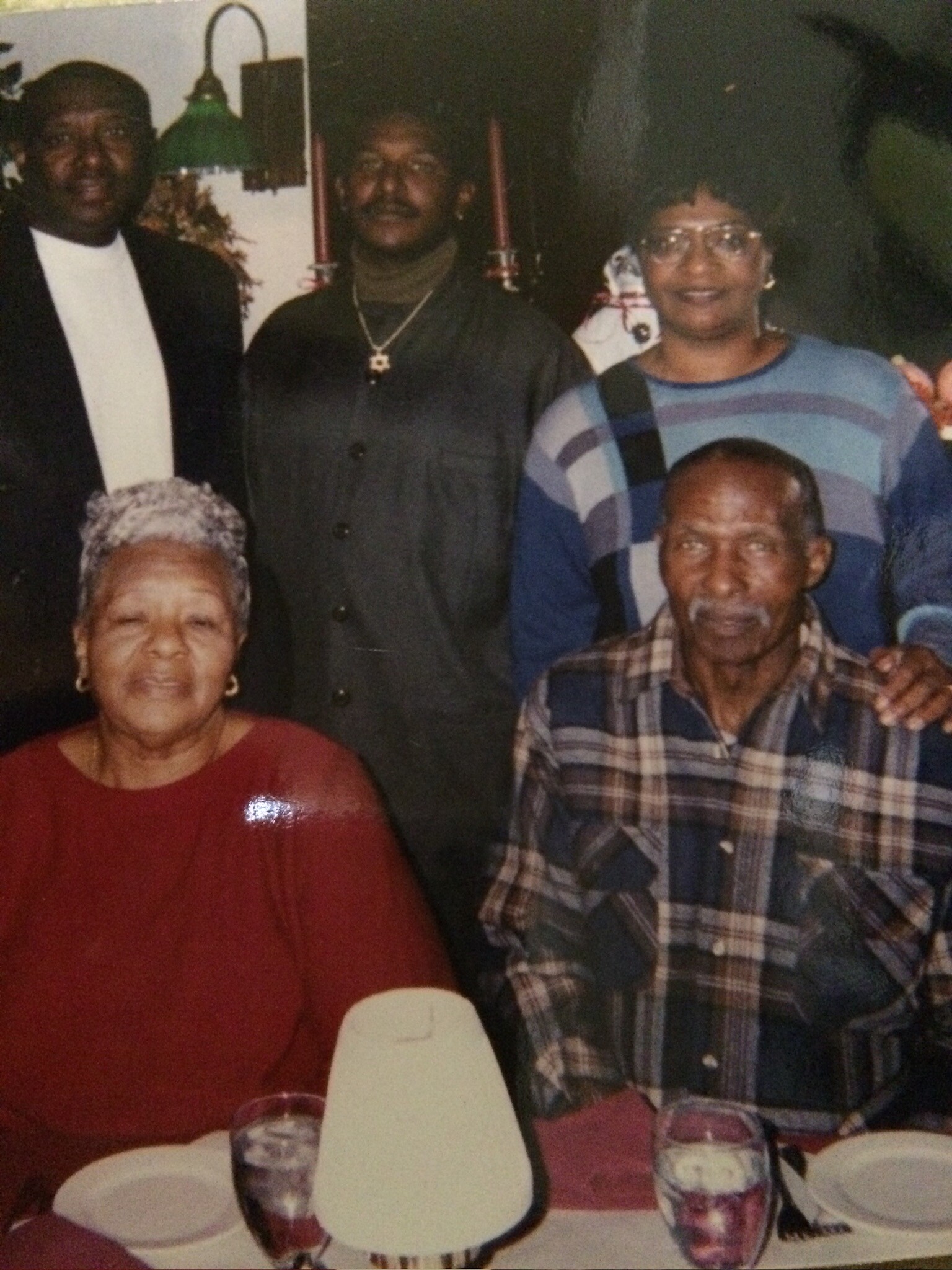 Group of African American seniors posing around a table at a family gathering; woman in red foreground left, man in plaid shirt foreground right, with three adults standing behind them.