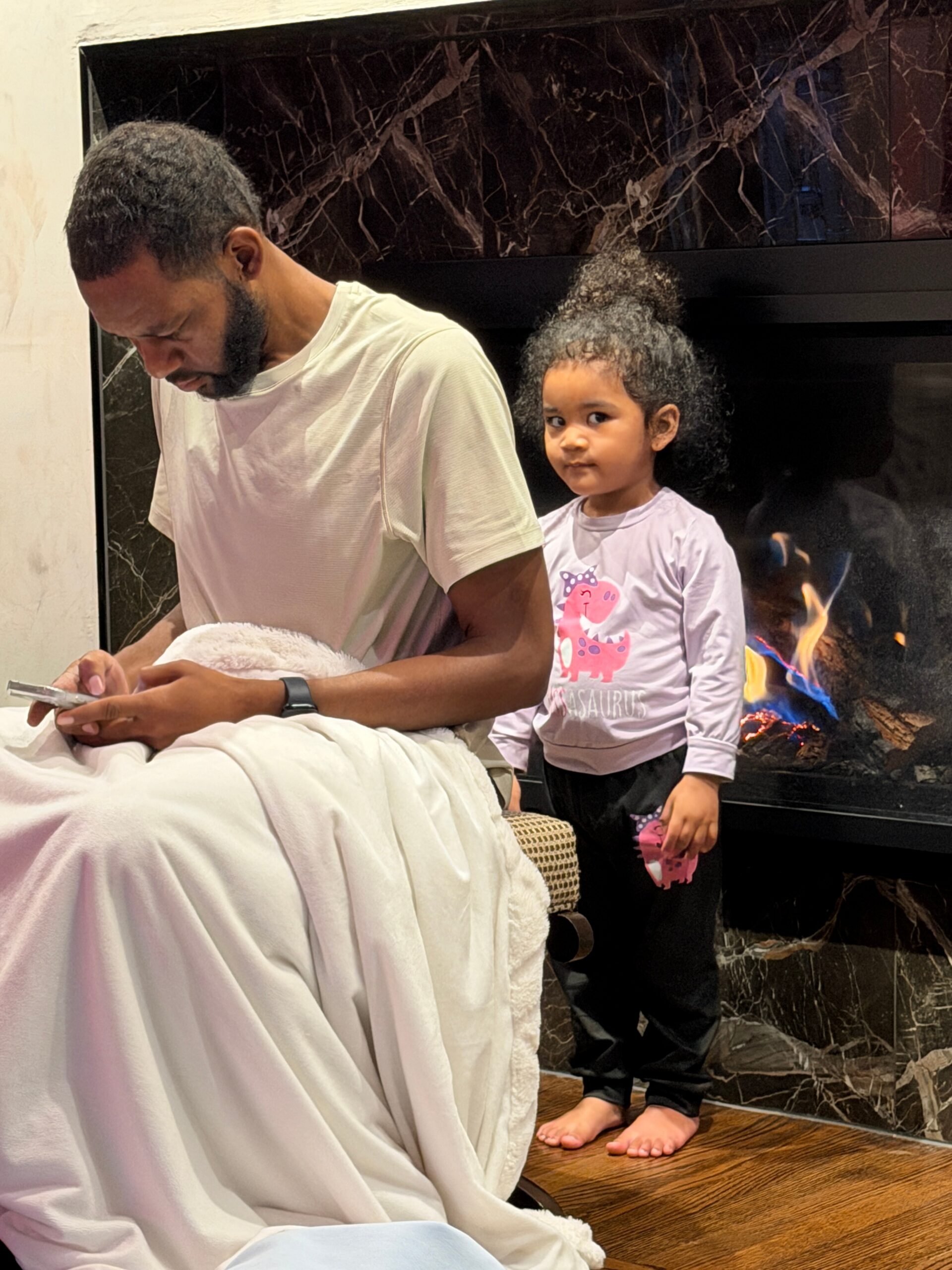 Man with short hair and beard sits on a bed covered with a white blanket, looking at a phone while a young girl stands behind him by a marble fireplace, watching.