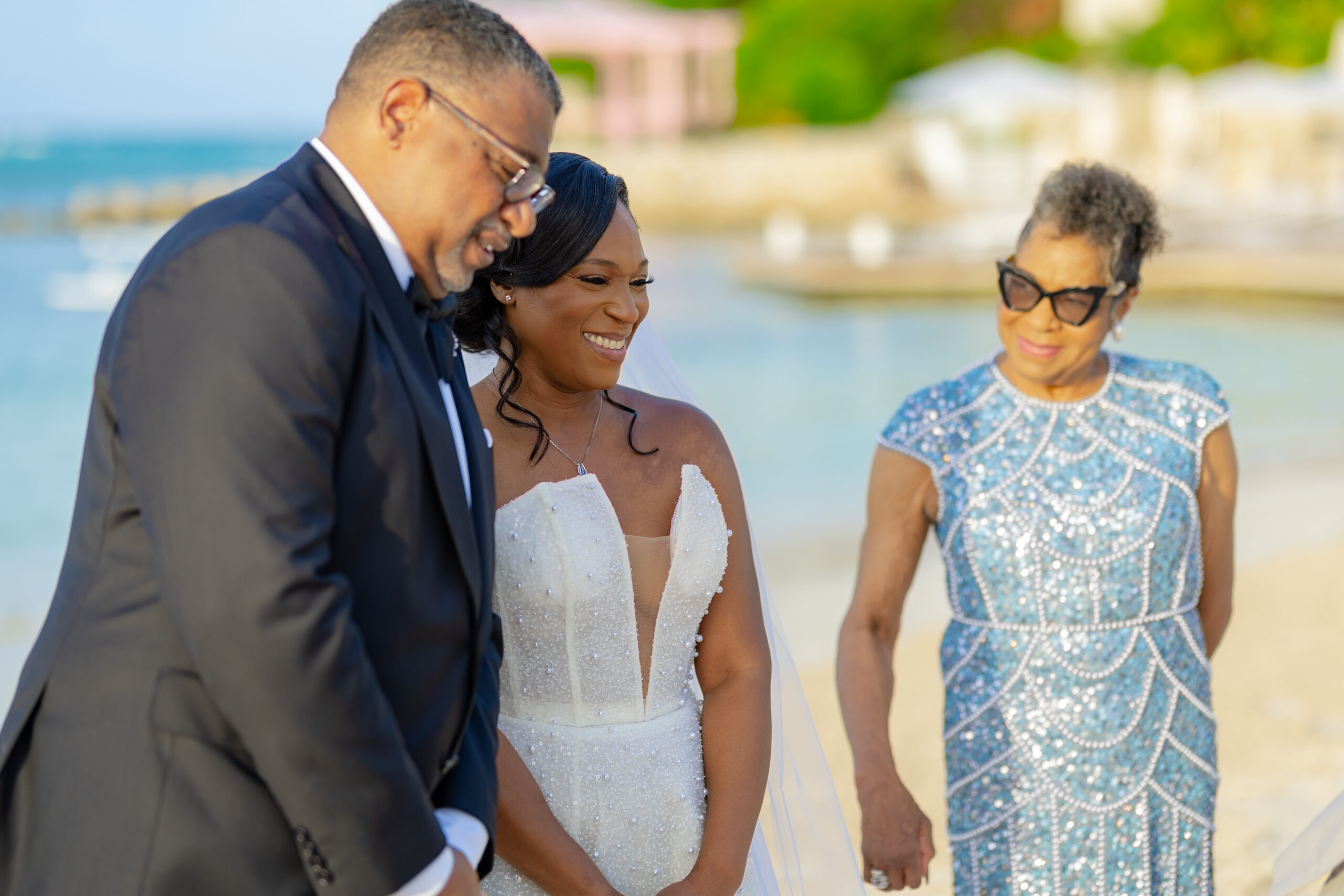 Bride in a white beaded wedding gown smiles as she stands with a man in a tuxedo and a woman in a sparkling blue dress on a sunny beach.