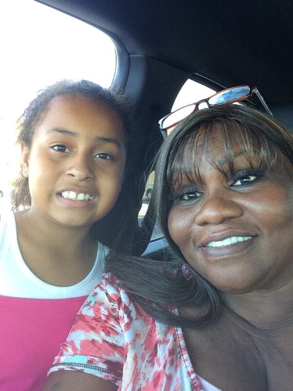 Two people smiling for a selfie in a car: a young girl and an older woman in the front seat, facing the camera.