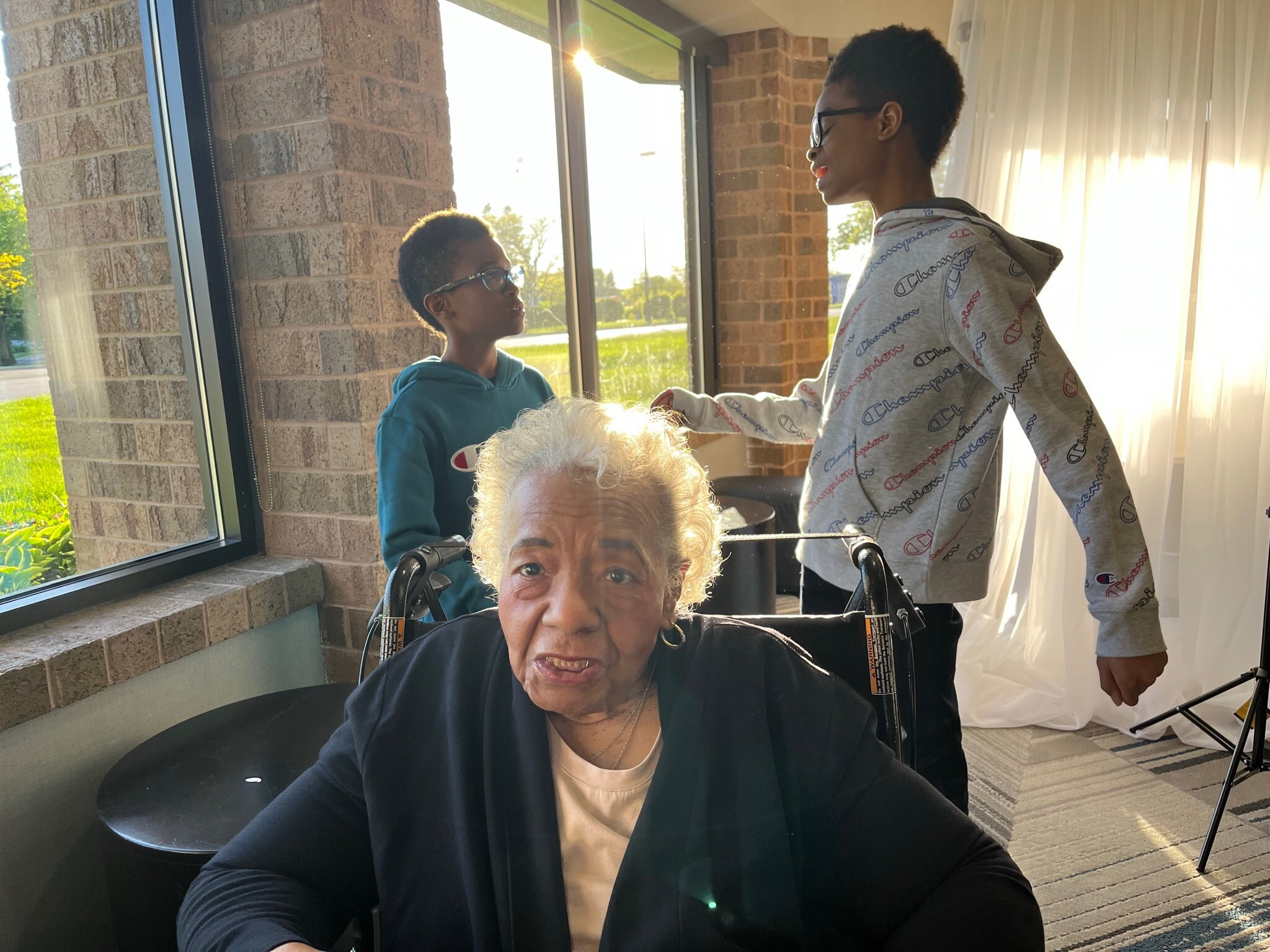 Elderly woman in a wheelchair at a lobby table, assisted by two younger people near sunlit windows.