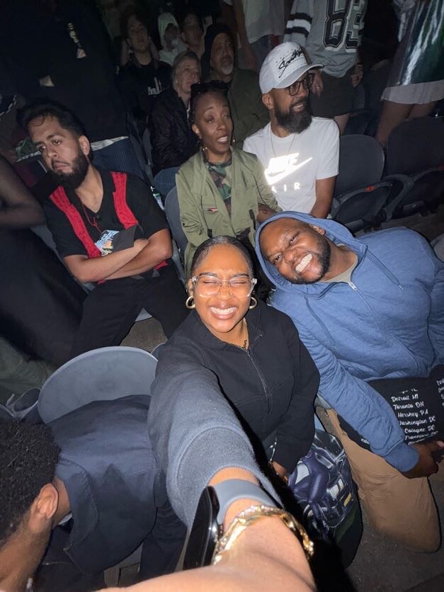 A group of friends at a crowded event, two people in front taking a selfie while others sit behind in bleacher seats.