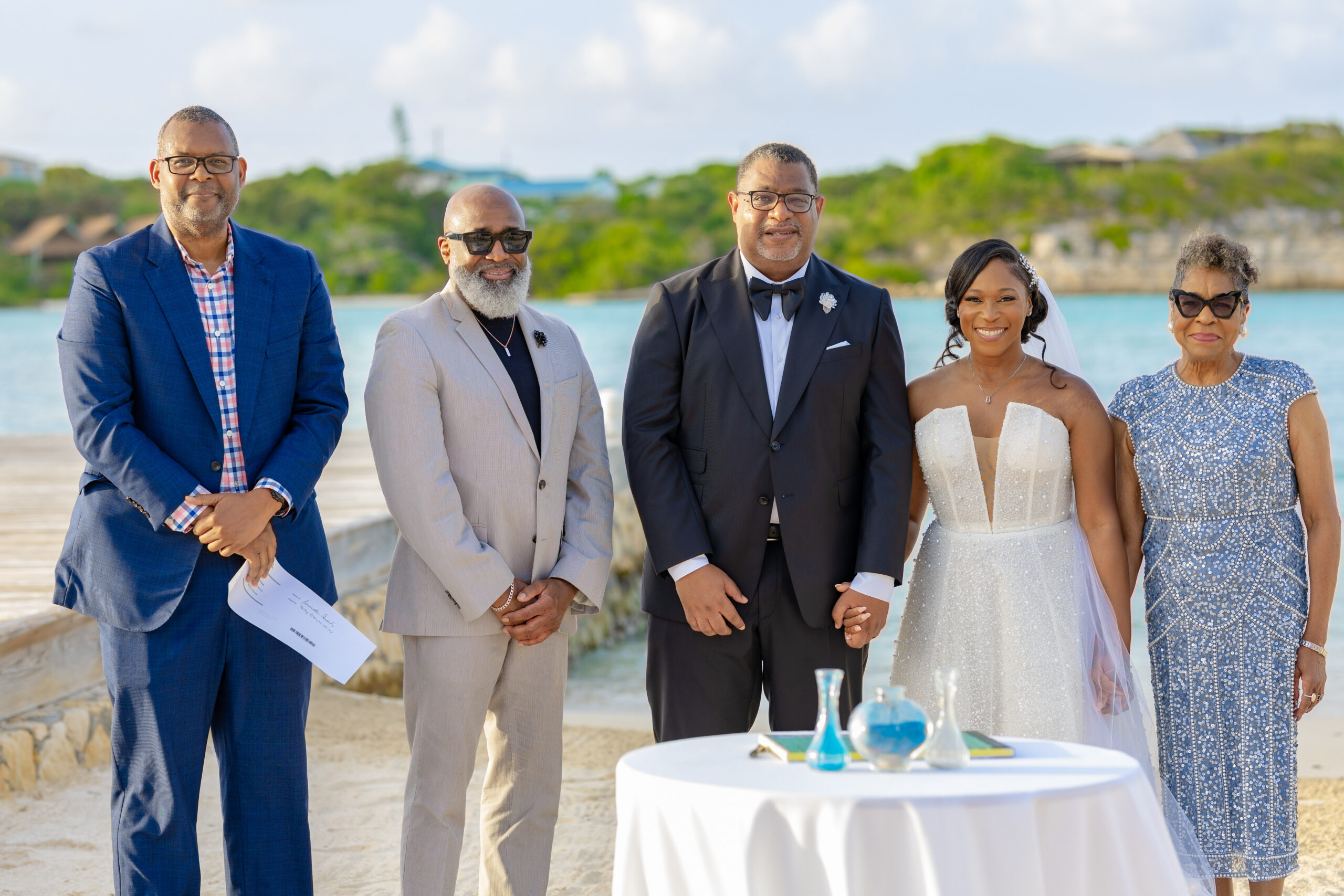 Group of five adults in formal attire posing at a seaside wedding with a table of drinks in the foreground.