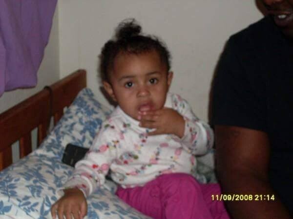 Toddler sitting on a bed wearing a white floral top and pink pants, finger near mouth, with an adult beside her.