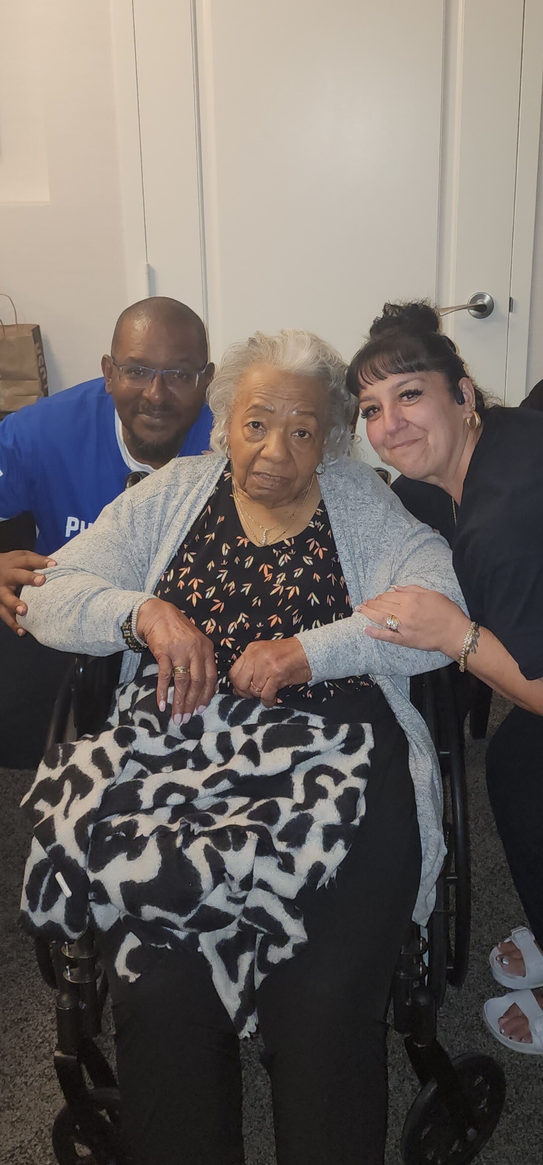 Three adults sit together indoors: an elderly woman in a wheelchair wrapped in a black-and-white blanket, flanked by a man and a woman smiling at the camera.