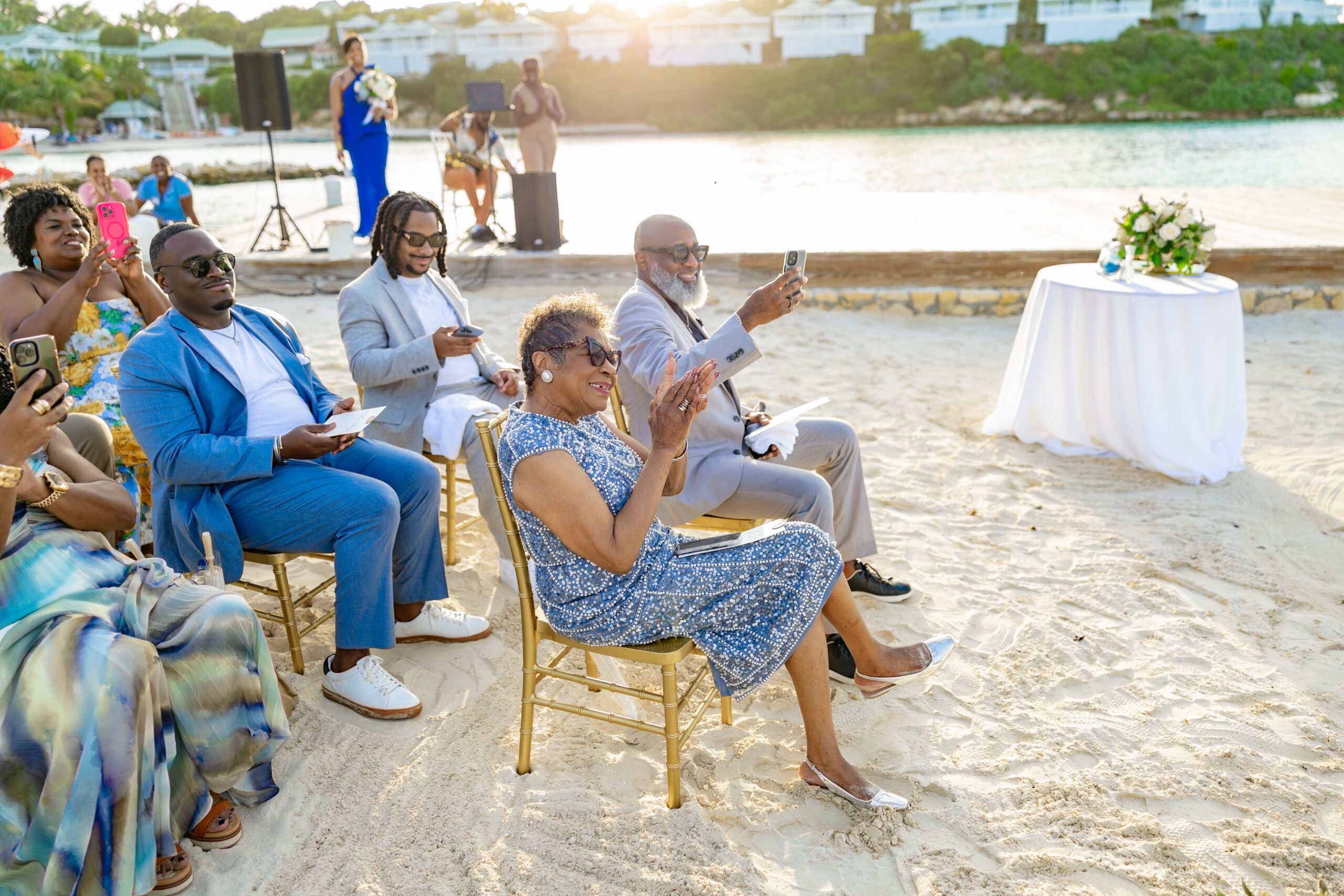 Group of guests seated on beach chairs at a sunny seaside ceremony, many taking photos with smartphones.