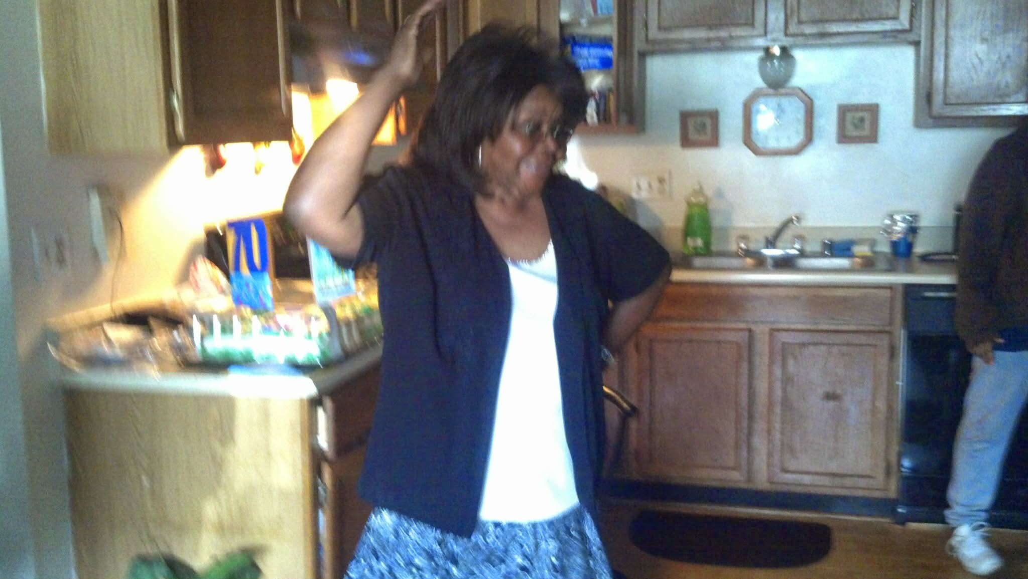 Woman in a home kitchen raising her right hand, smiling, wearing a dark blazer over a white top and a patterned skirt.