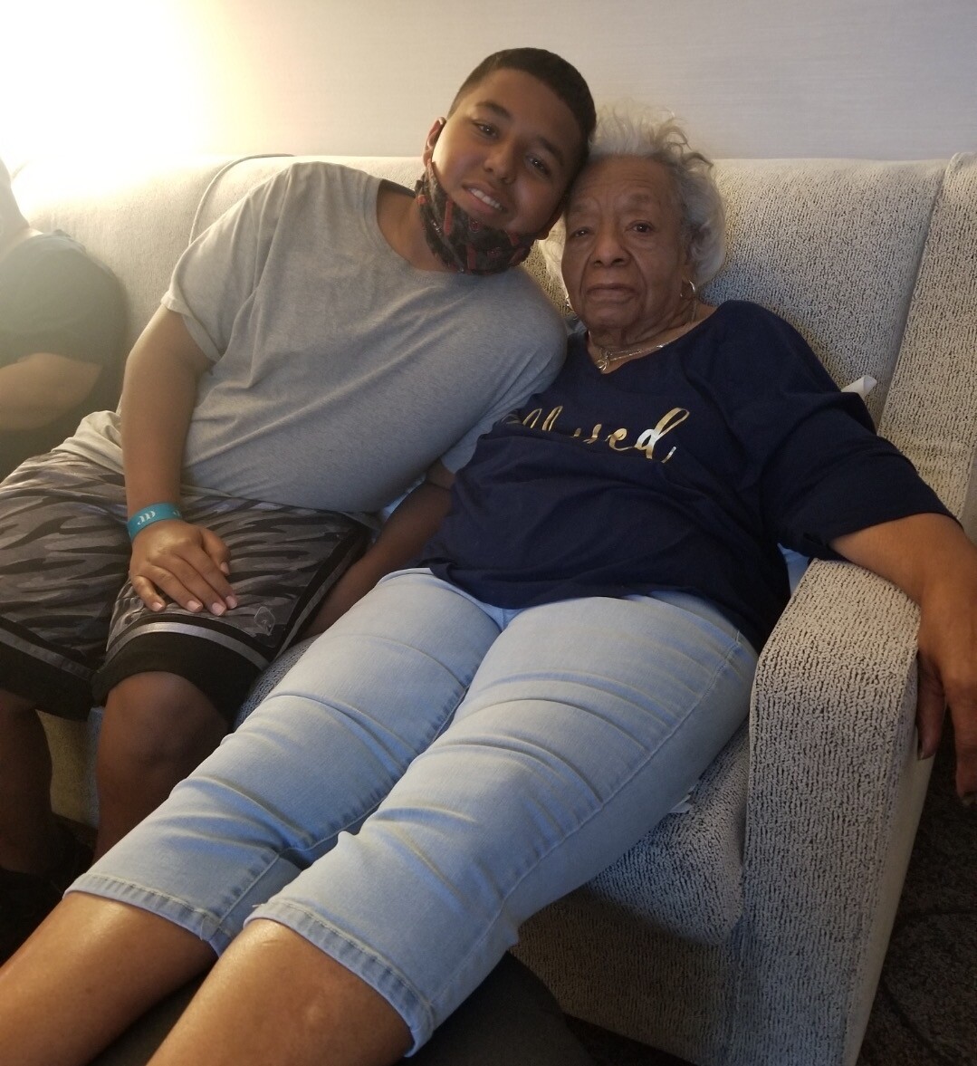 Young man and elderly woman sitting close together on a beige couch, smiling at the camera.