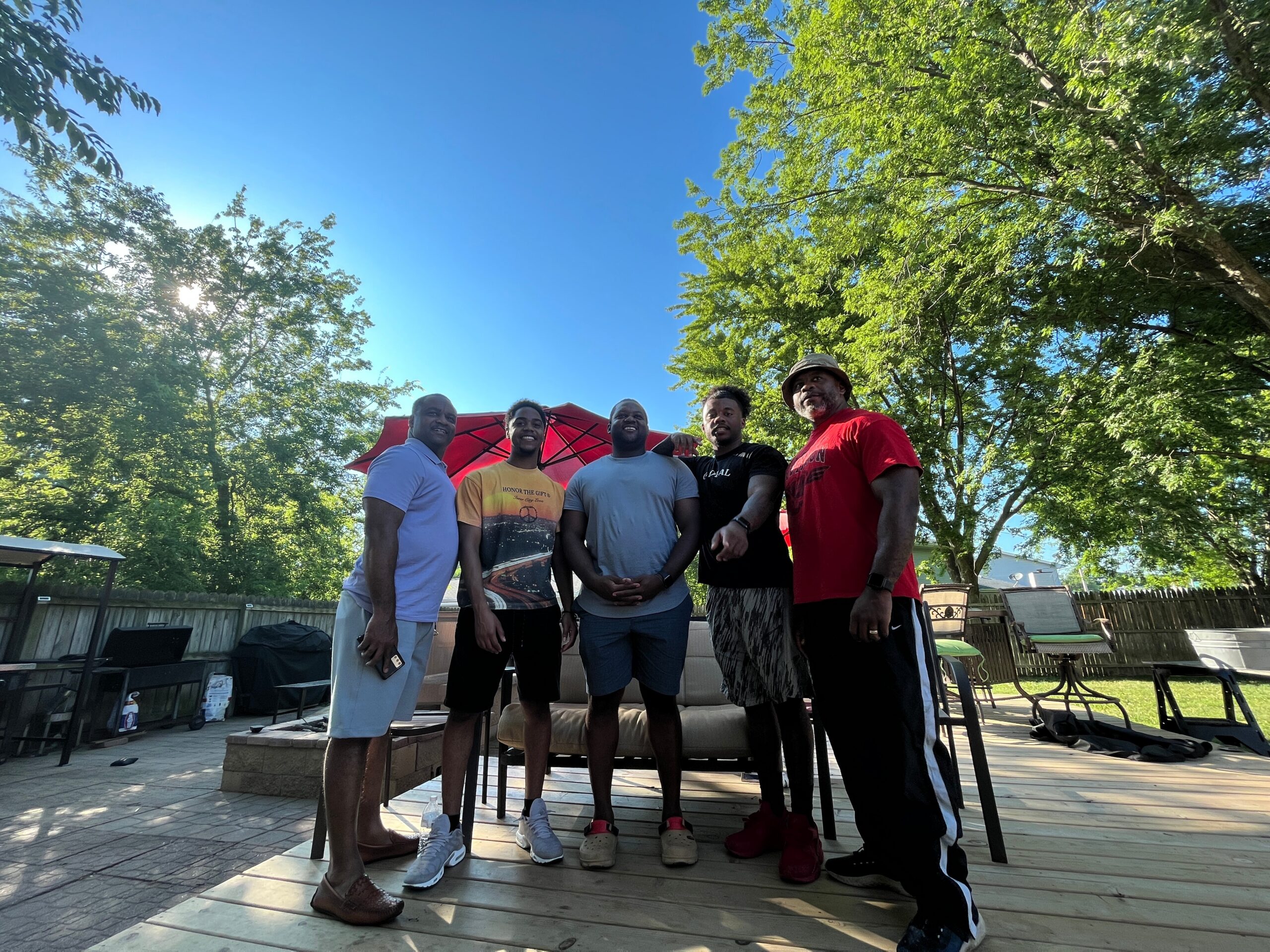 Six adults standing together on a backyard patio under a bright blue sky, with a red umbrella and green trees in the background.
