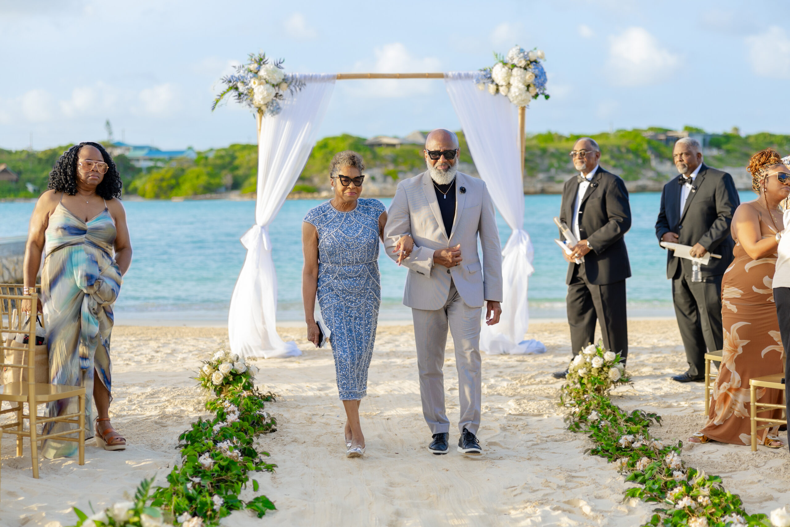 Couple and guests walk toward a white-draped seaside wedding arch on the beach with floral decorations.