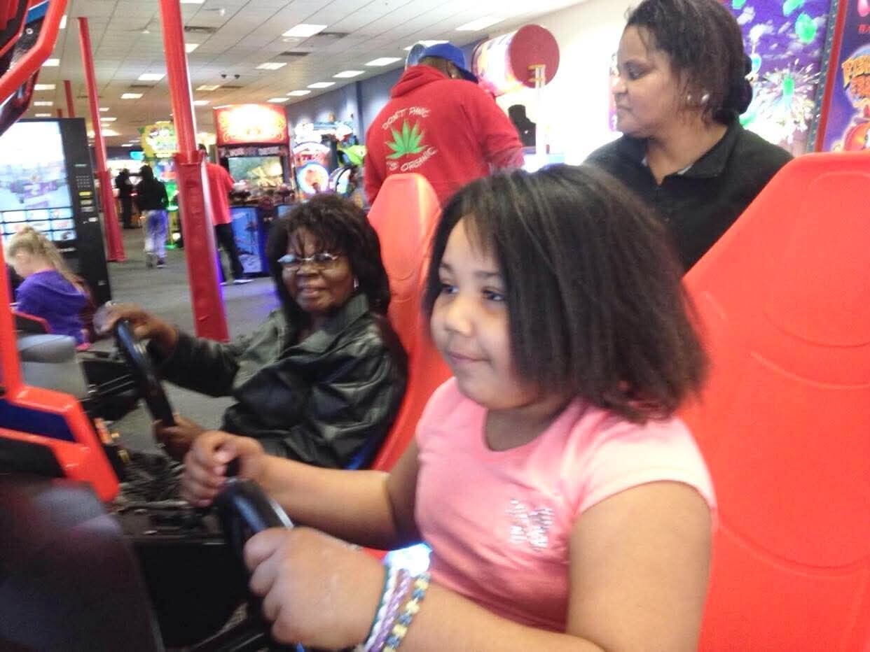 Young girl in a pink shirt sits in a red arcade ride car, smiling, while an adult woman sits beside her and another person stands behind in a busy arcade.